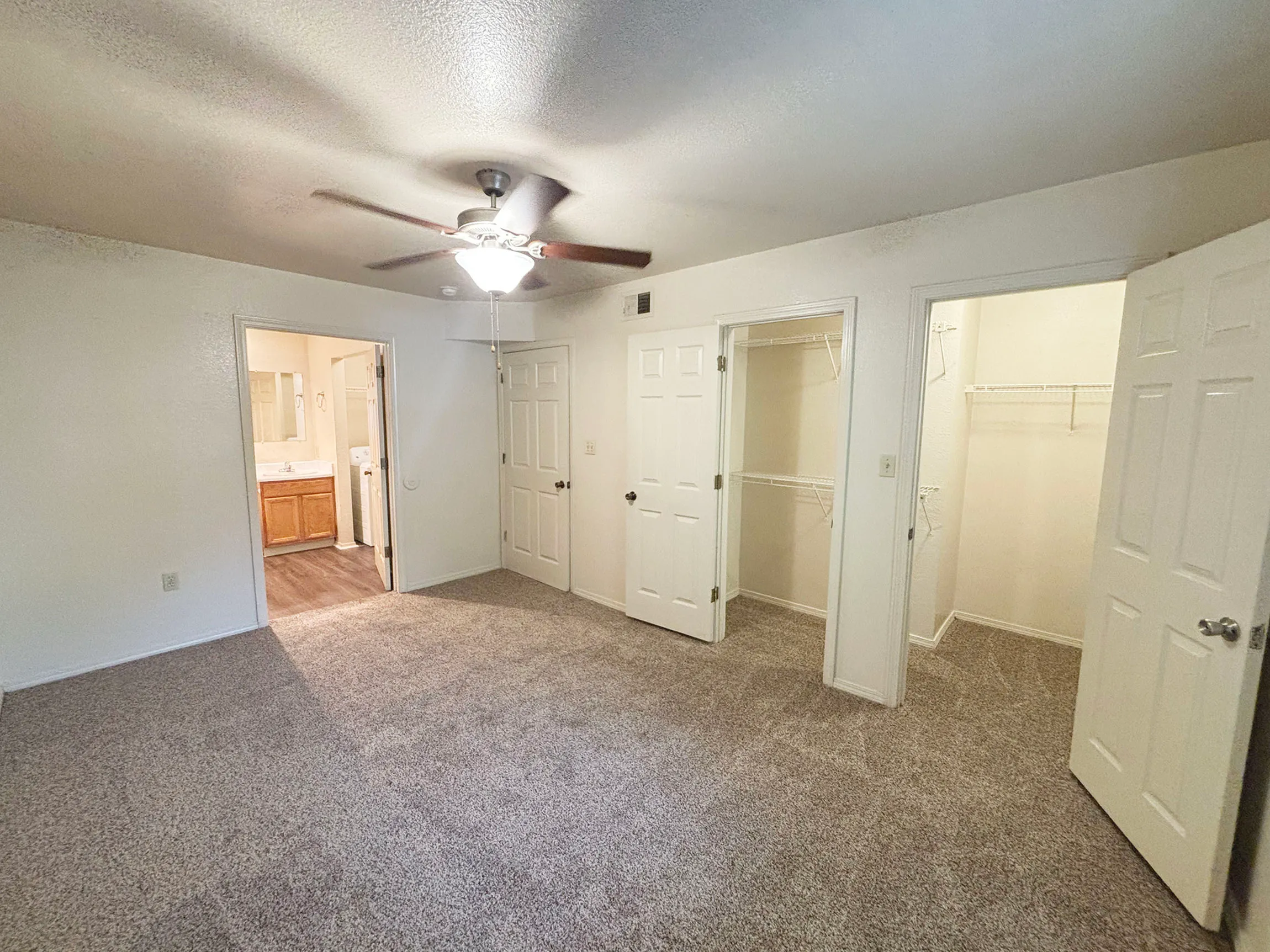 Spacious bedroom with plush carpeting, ceiling fan, and dual walk-in closets adjacent to an elegant bathroom.