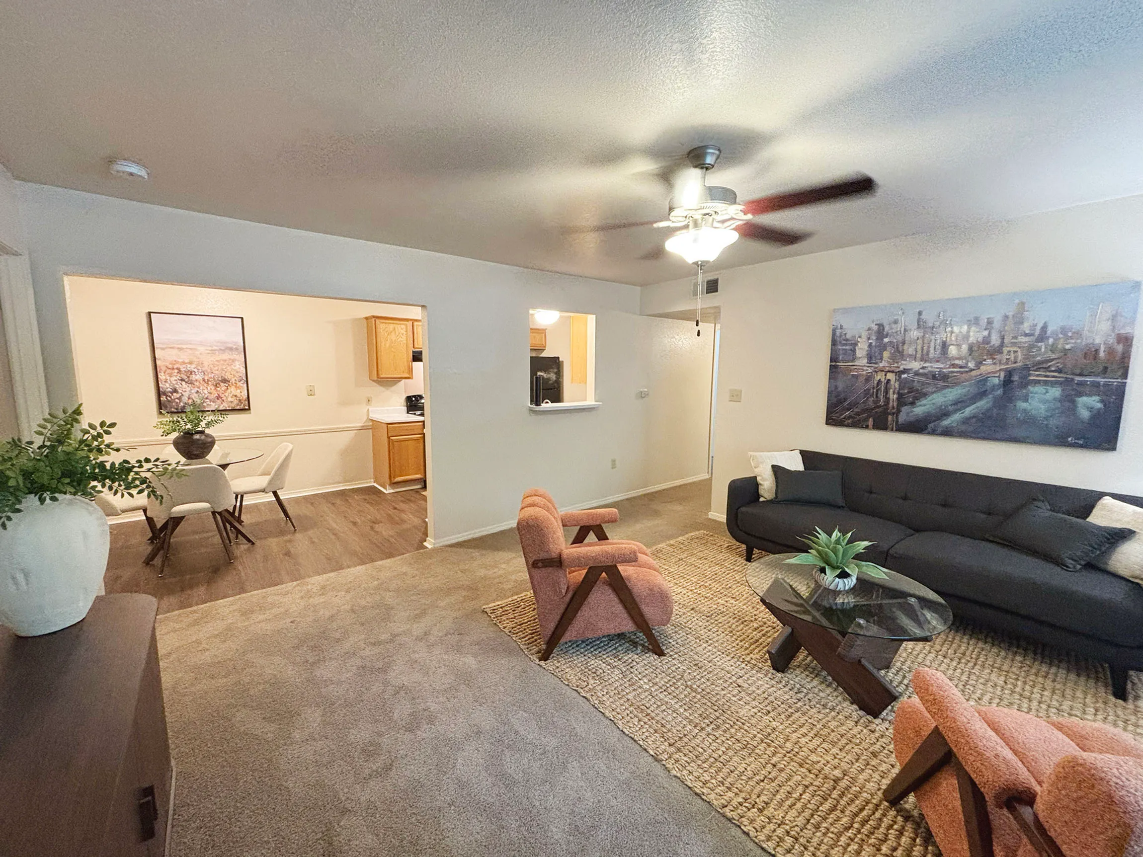 Cozy living room with modern decor, featuring a stylish glass coffee table, inviting gray sofa, and vibrant artwork. Adjacent dining area with elegant seating provides a seamless flow into the kitchen.