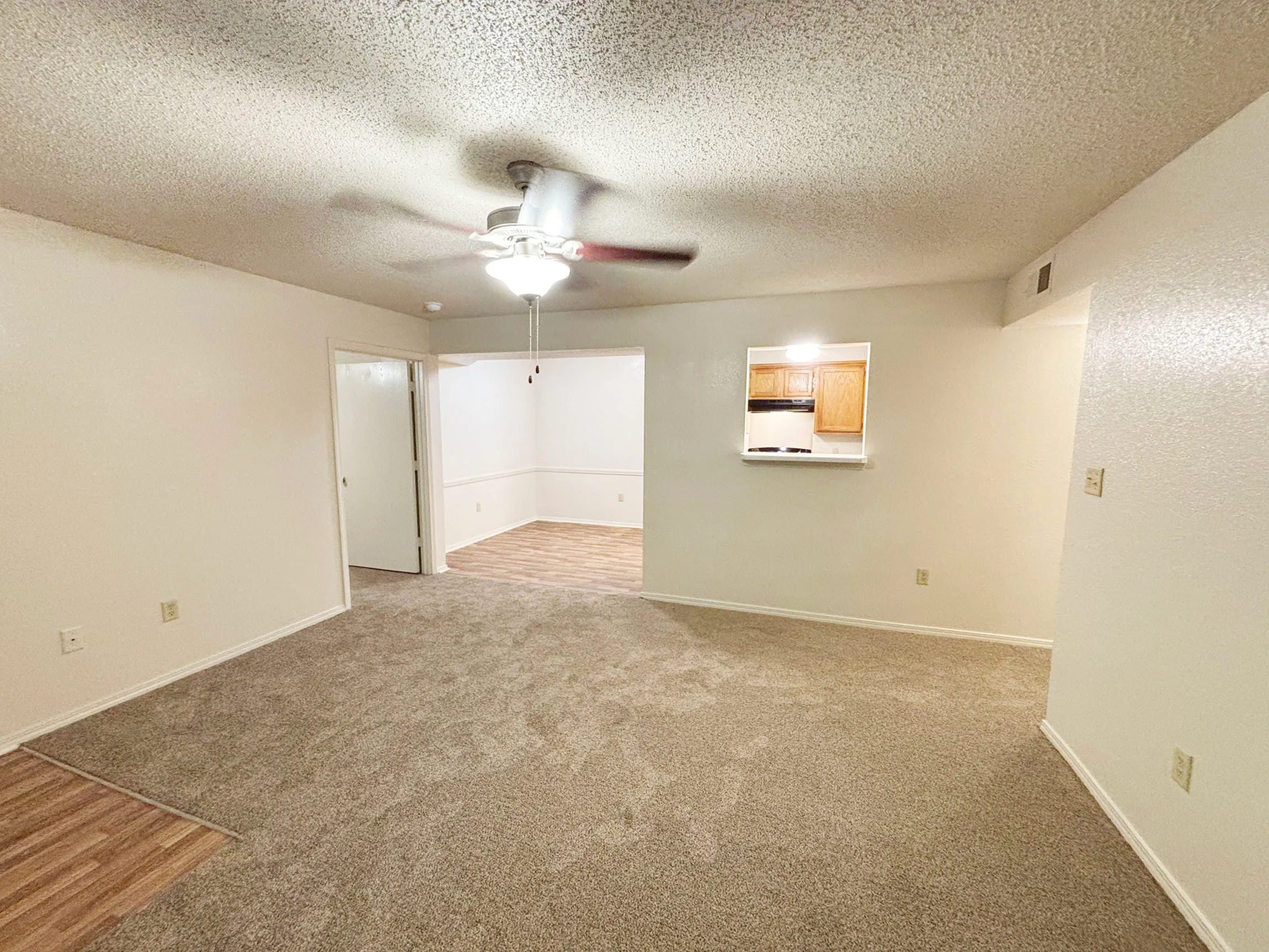 Inviting living space with plush carpeting, modern ceiling fan, and a view into the cozy kitchen with wooden cabinetry.