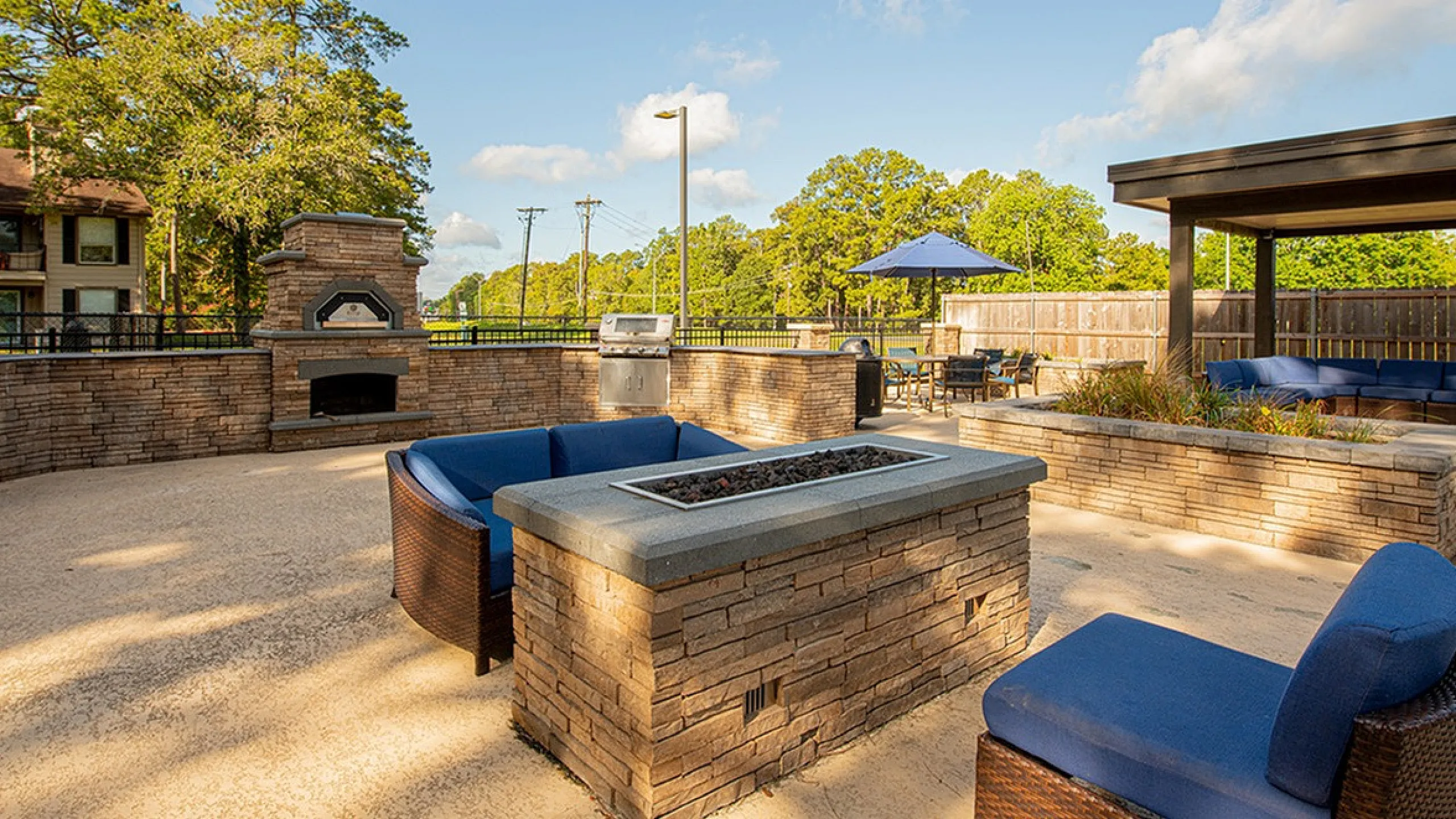 Charming outdoor patio with cozy blue seating, a stylish fire pit, and a brick pizza oven, perfect for entertaining under a clear sky.
