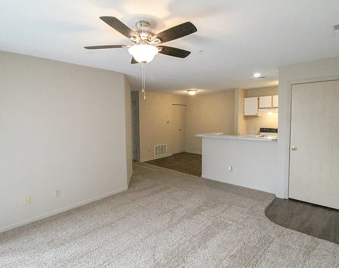 Bright and welcoming living area with plush carpeting, ceiling fan, and adjacent modern kitchen space.