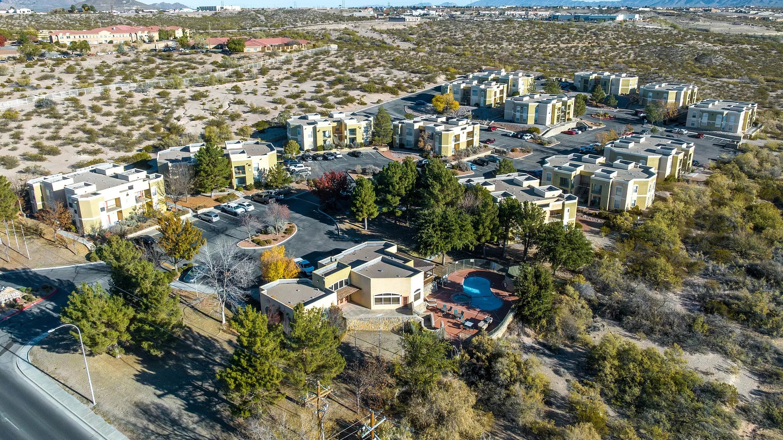 Scenic aerial view of modern apartment complex with lush greenery, inviting pool, and spacious parking, nestled within a serene desert landscape.