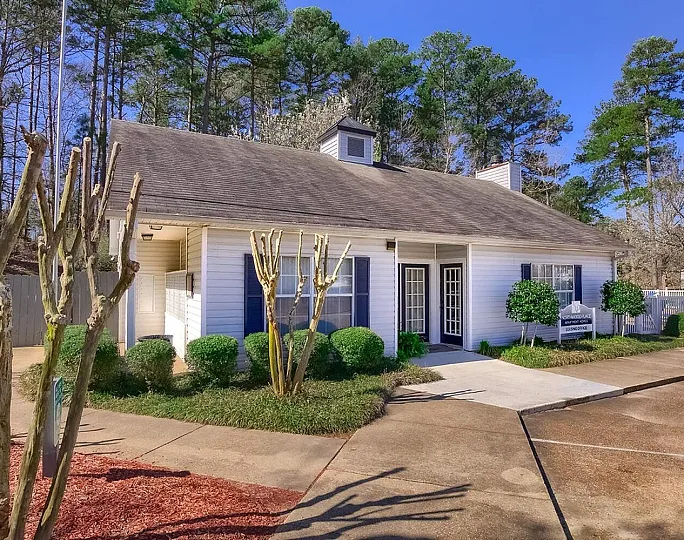 Inviting leasing office surrounded by lush greenery and serene wooded area, showcasing charming white facade and welcoming entry pathway.