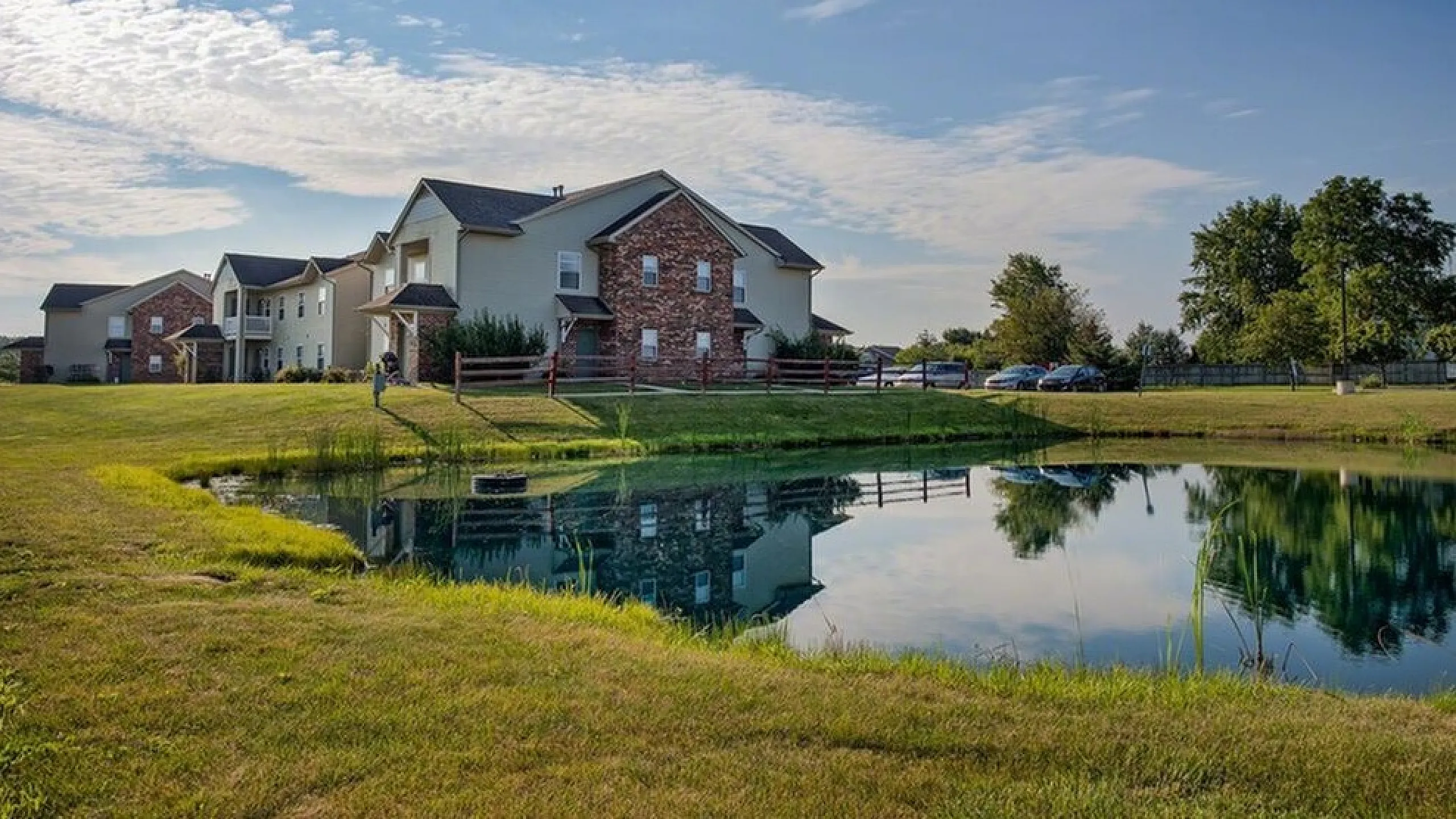 Charming apartment buildings with classic architectural design nestled beside a tranquil pond, surrounded by lush greenery and a clear blue sky.