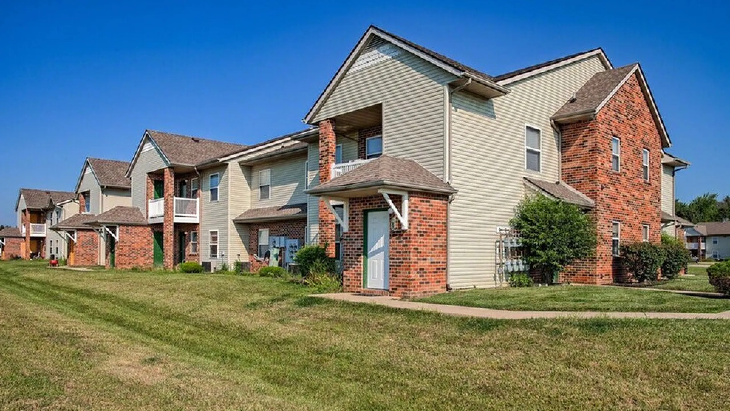 Charming suburban apartment complex with modern brick and wood exterior, featuring well-kept lawns and inviting pathway in a serene neighborhood.