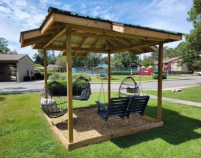 Charming outdoor sitting area with cozy hanging chairs and a swing set under a wooden pergola, surrounded by lush greenery and a vibrant playground in the background.