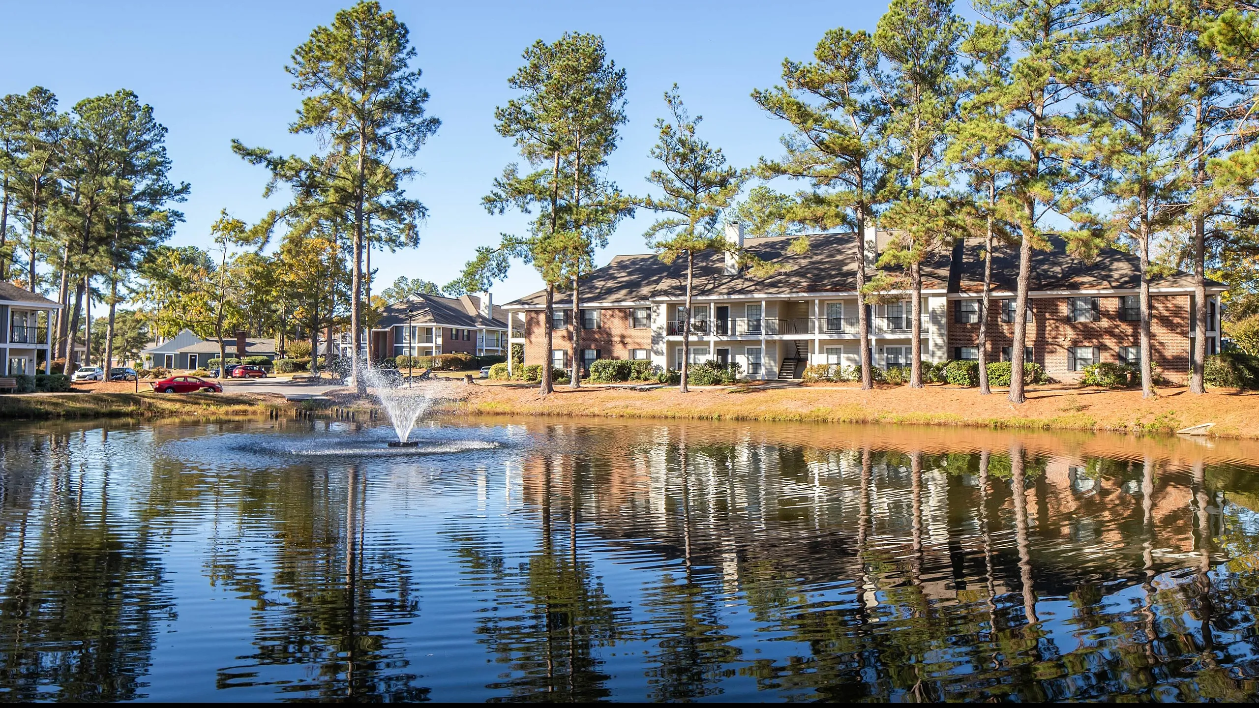 Serene apartment complex with scenic pond, fountain, and majestic pine trees under clear blue skies.