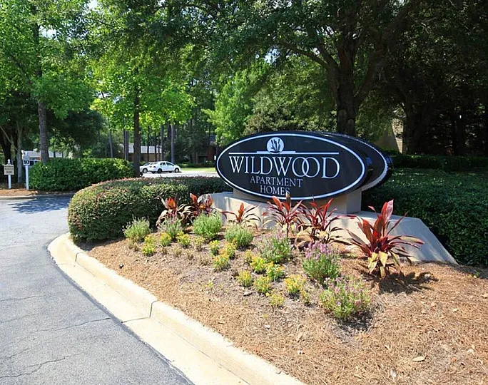 Inviting entrance to Wildwood Apartment Homes surrounded by lush greenery, vibrant plants, and scenic landscaping.