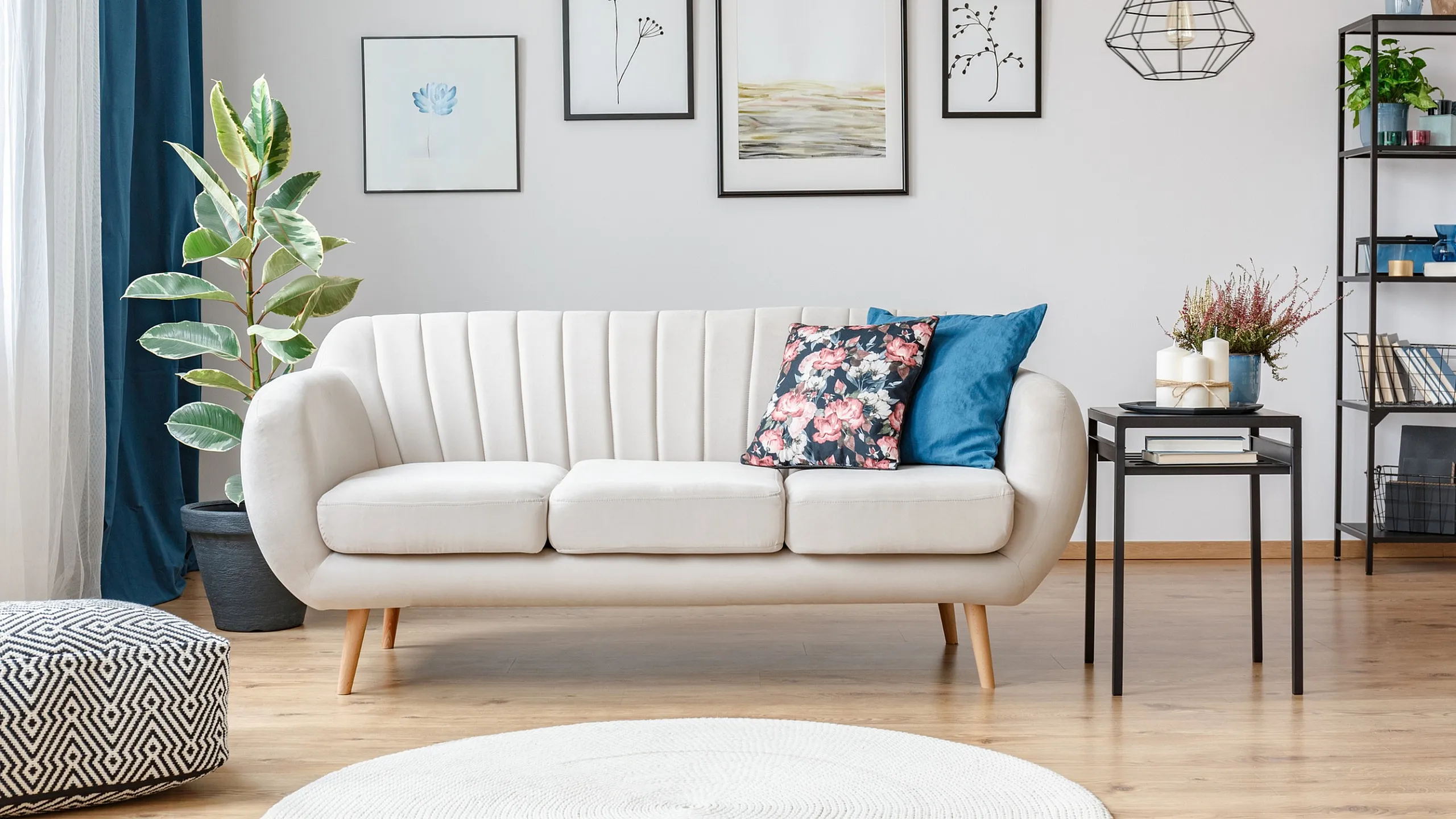 Chic living room with modern white sofa, floral and blue cushions, artistic wall decor, elegant lighting, and lush green plant accents.