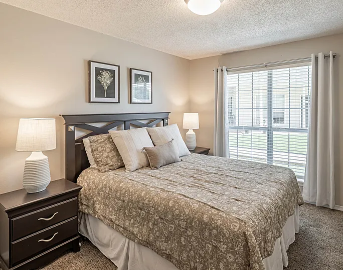 Bright and cozy bedroom featuring a plush bed with elegant bedding, soft lighting, and large window with white curtains offering natural light.
