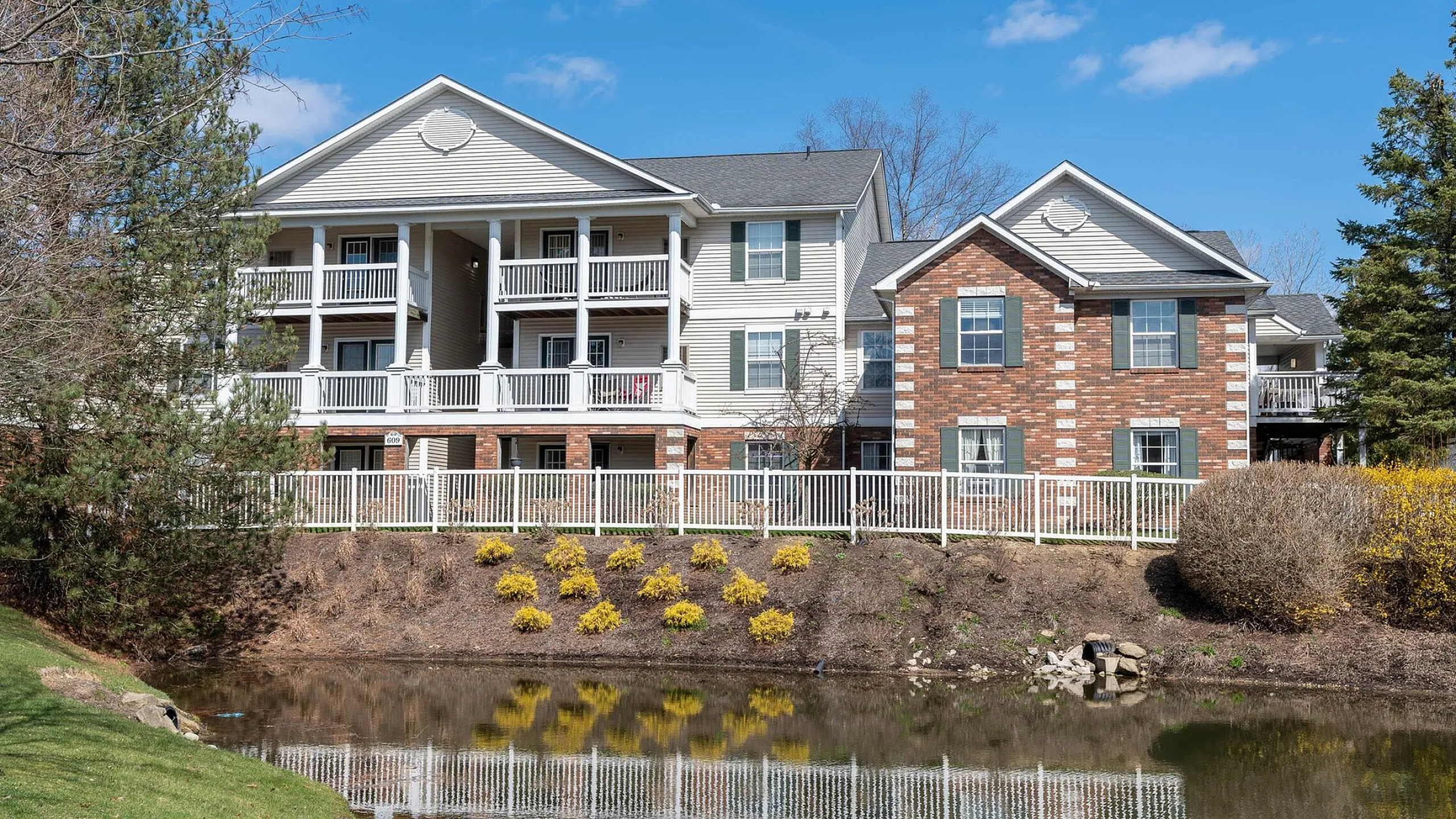 Charming apartment building with spacious balconies overlooking a serene pond, surrounded by lush greenery and vibrant landscaping.