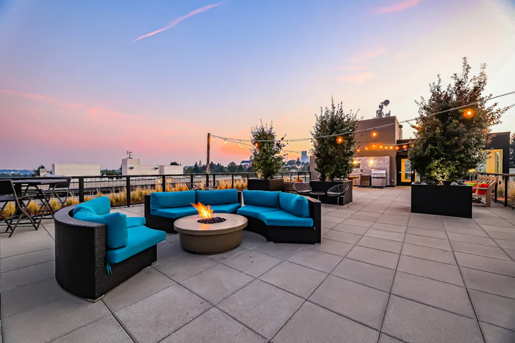 Chic rooftop terrace with cozy fire pit seating, vibrant blue cushions, and ambient string lighting at sunset overlooking cityscape.