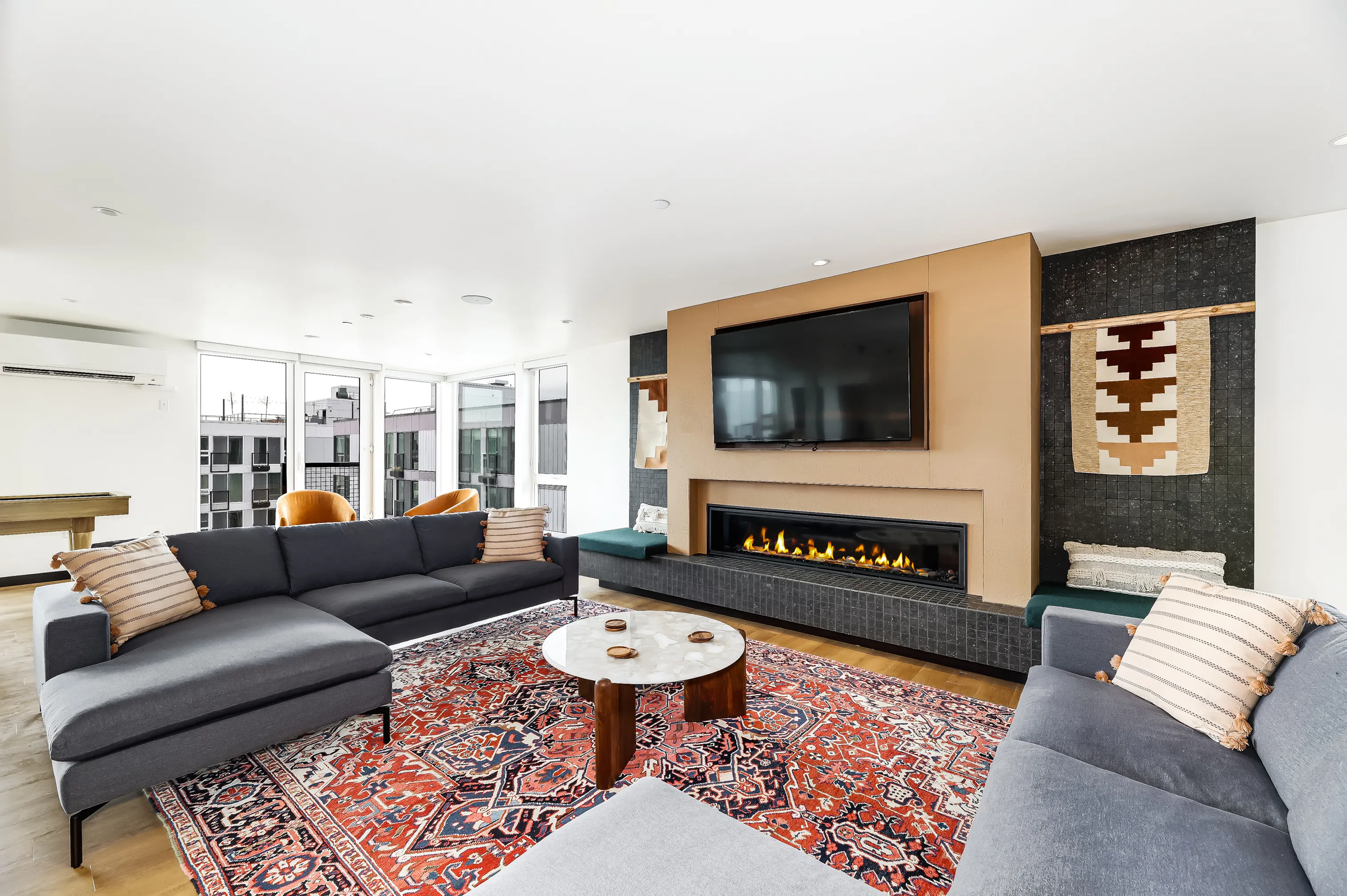 Luxurious living room boasting modern gray sofas, vibrant area rug, and a sleek fireplace beneath a large wall-mounted TV, complemented by floor-to-ceiling windows and stylish decor.