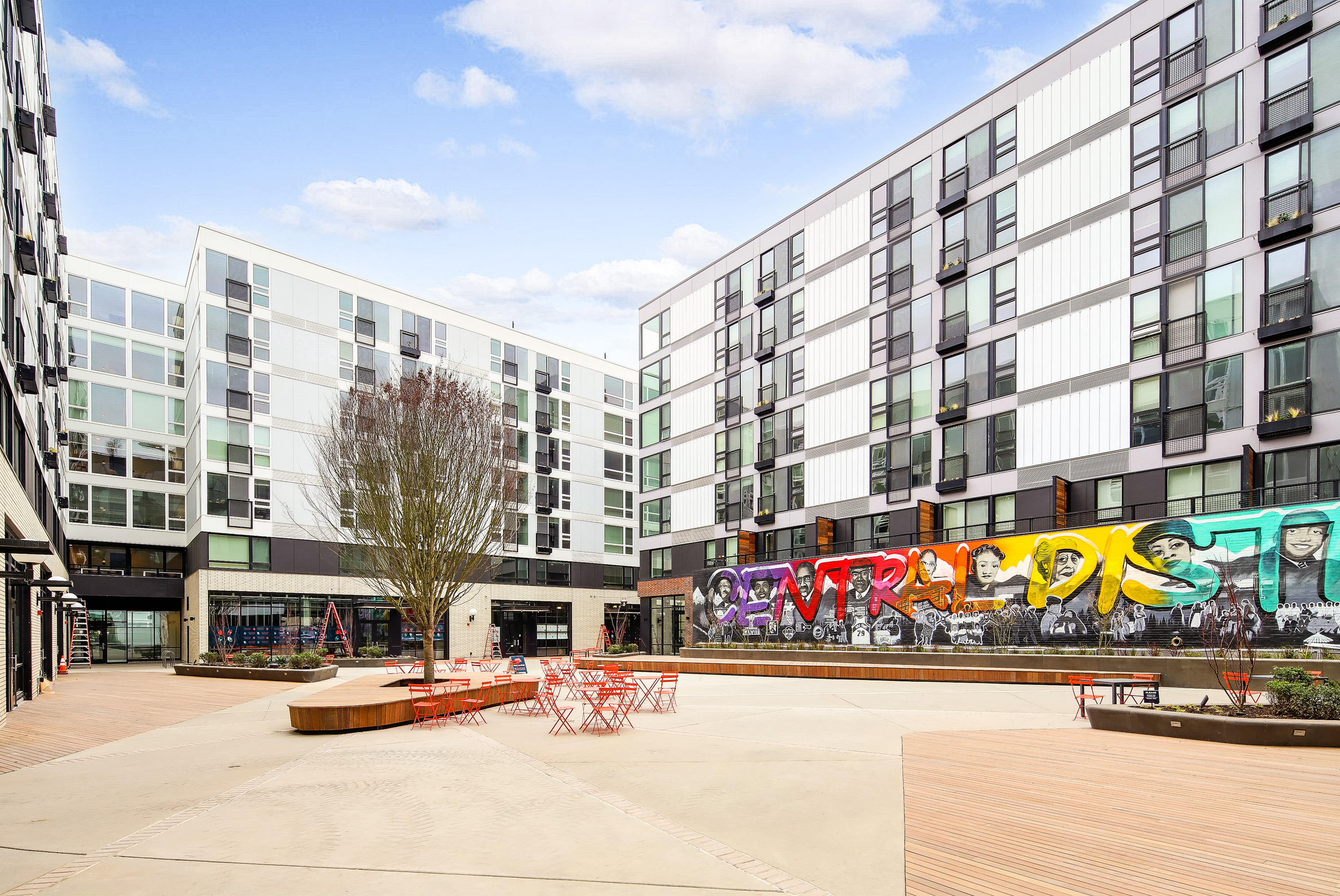 Vibrant apartment complex exterior with modern architecture and large colorful mural, featuring open outdoor seating and lush greenery.