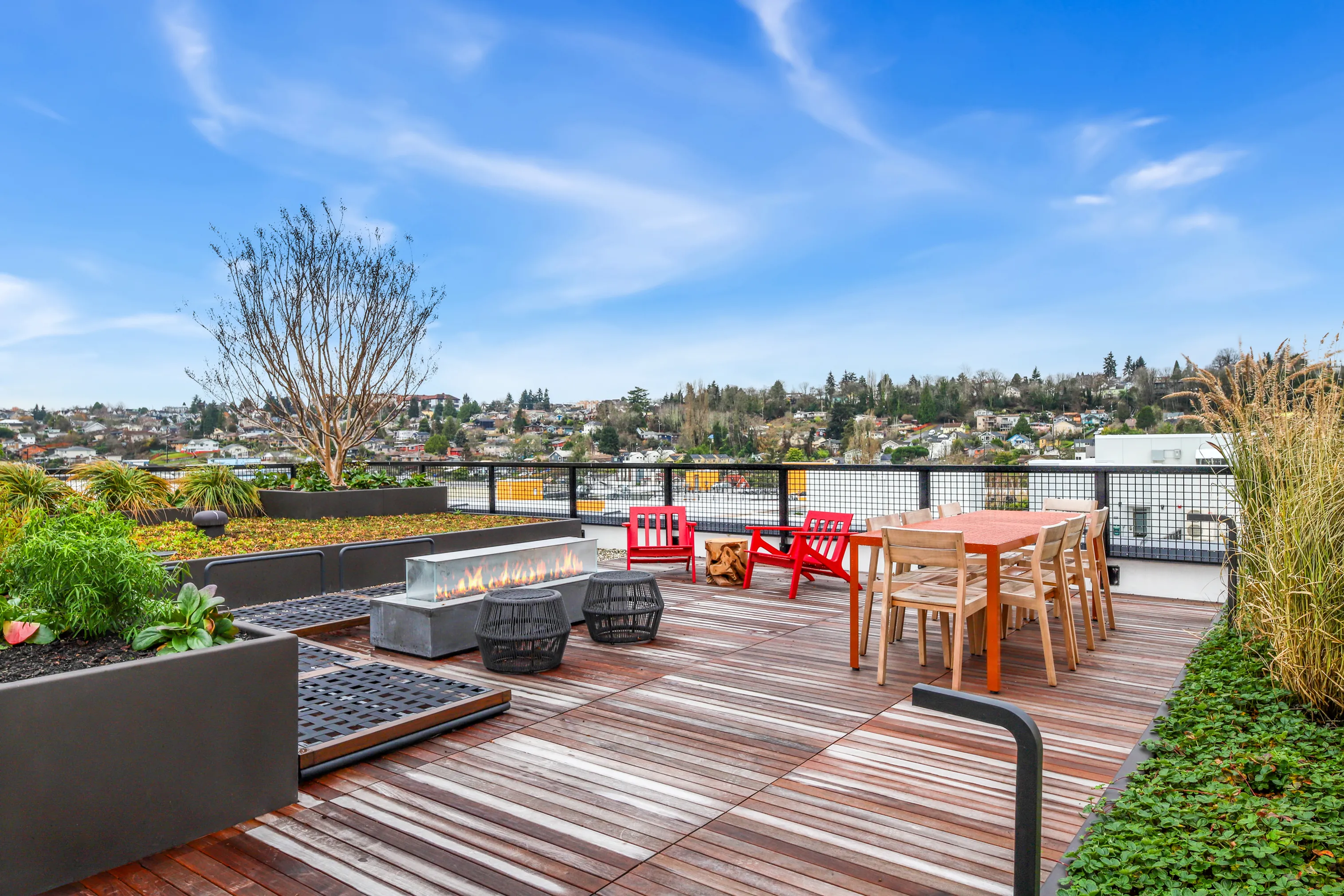 Elegant rooftop terrace with modern fire pit, vibrant seating area, and lush greenery, offering stunning panoramic city views under a clear blue sky.