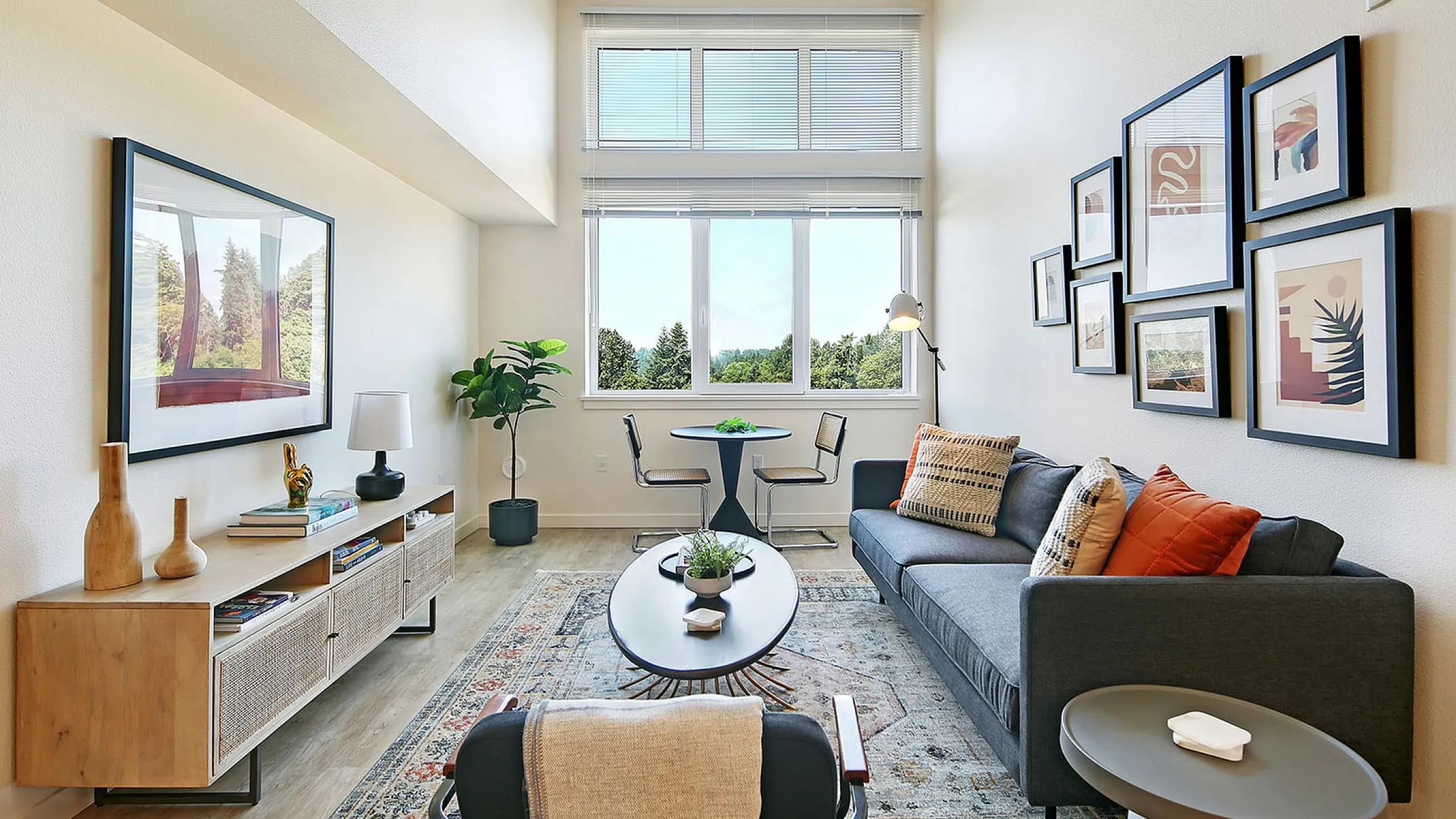 Bright and inviting living room featuring a stylish gray sofa, modern artwork, and a large window with lush green views.