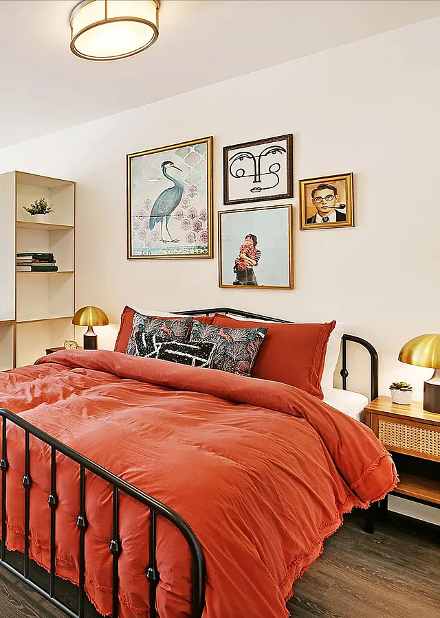Bright, modern studio apartment with a cozy red bed, stylish wall art, and a sunlit balcony overlooking greenery.