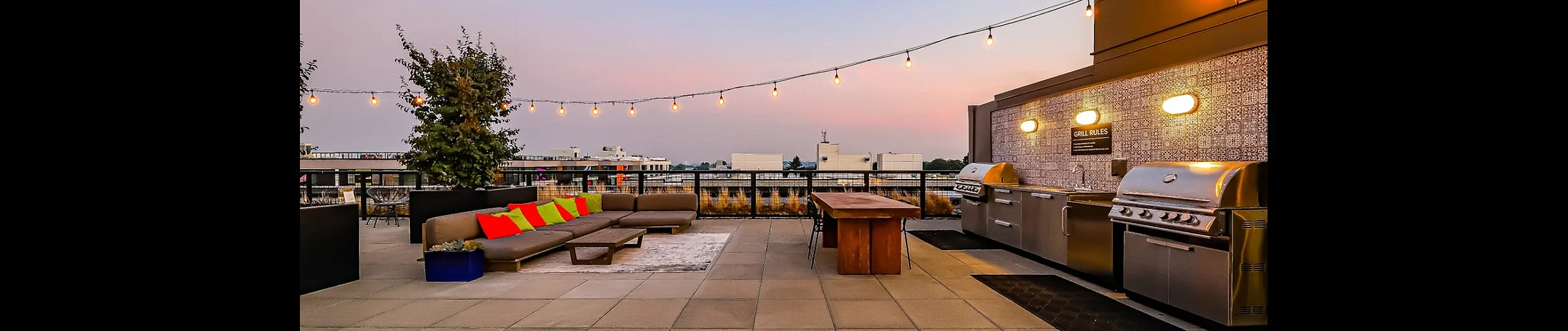 Chic rooftop patio with modern grill station, comfortable seating, and twinkling string lights against a picturesque sunset view.