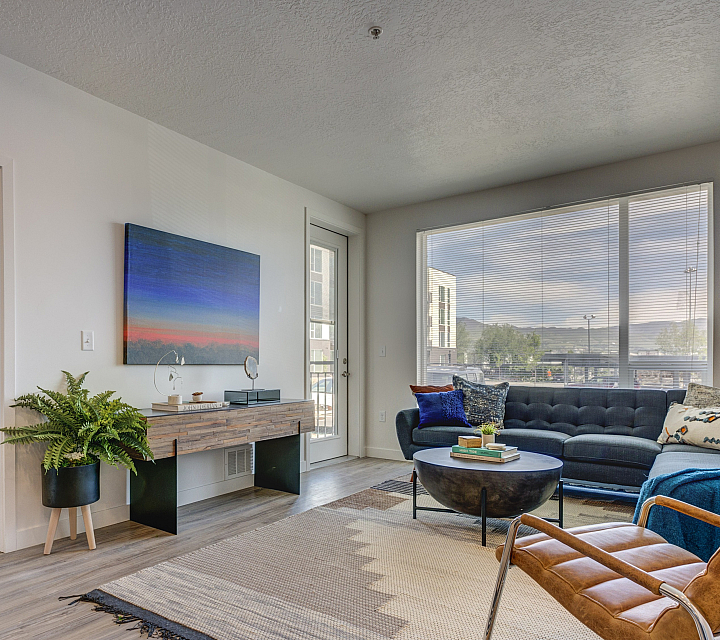 Bright and modern living room with large windows, featuring a cozy sectional sofa, stylish leather chair, and a glimpse into the inviting bedroom with contemporary decor.