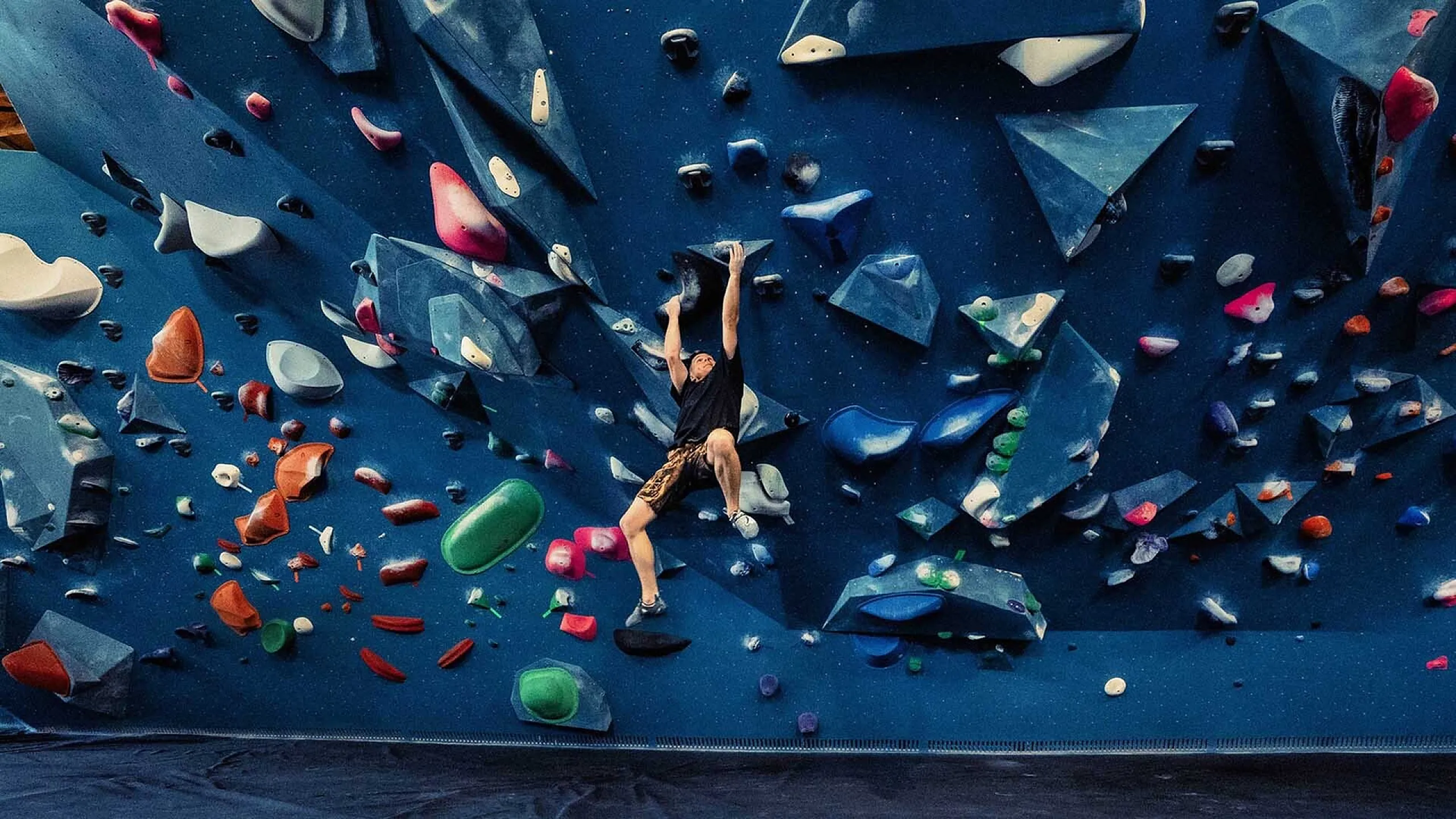 Dynamic indoor climbing wall with vibrant multi-colored holds and an athletic climber in mid-ascent, showcasing strength and agility.