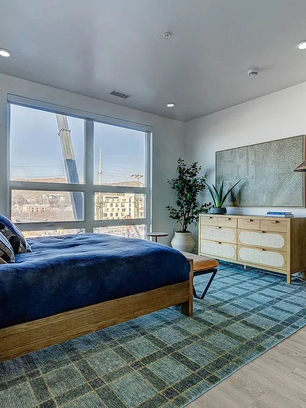 Modern apartment bedroom with large window, elegant decor, and cozy bedding, accompanied by stylish furniture and a contemporary lamp.