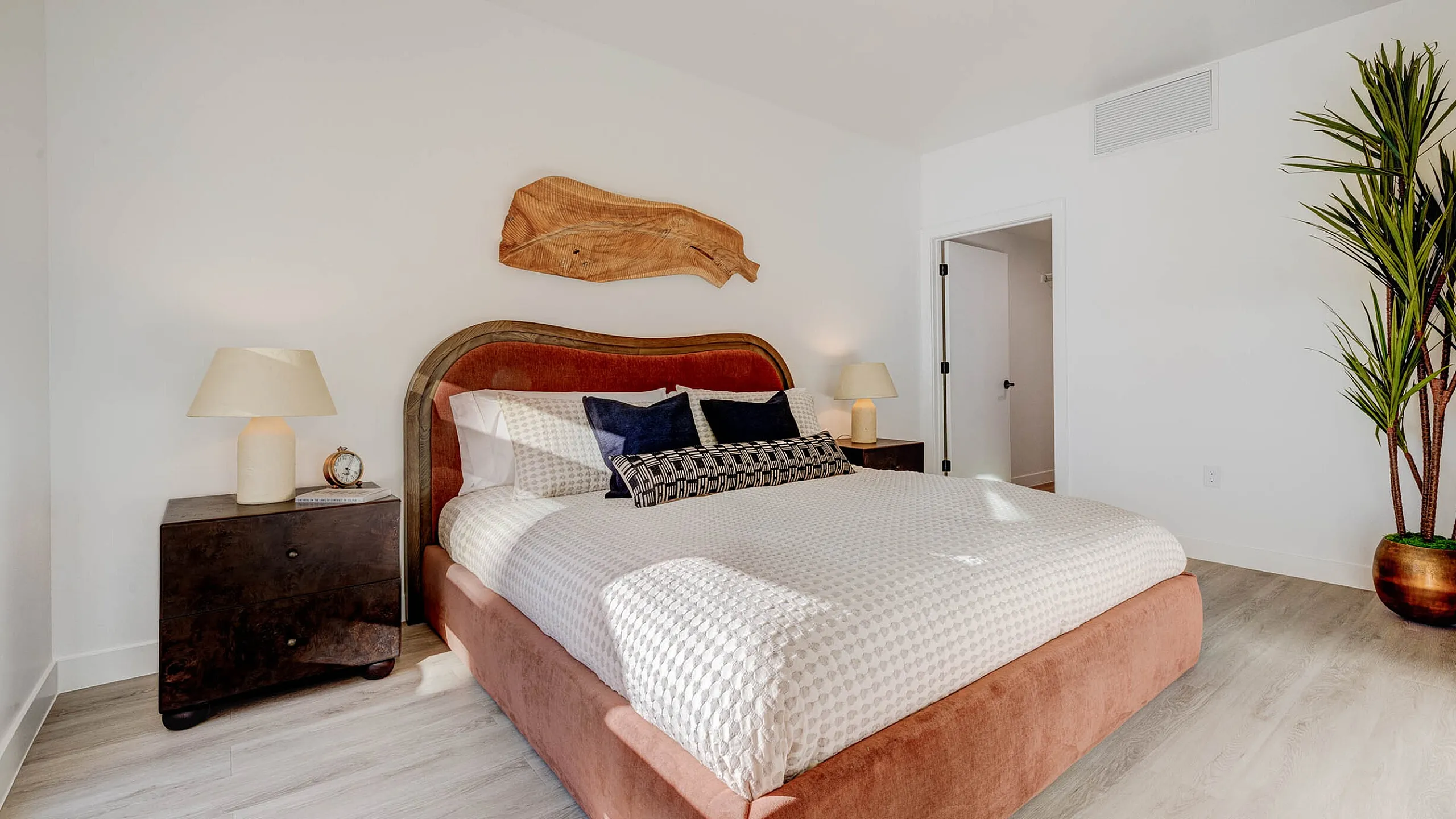 Elegant bedroom with a plush bed, stylish decor, and abundant natural light, complemented by a chic wooden sculpture and greenery.