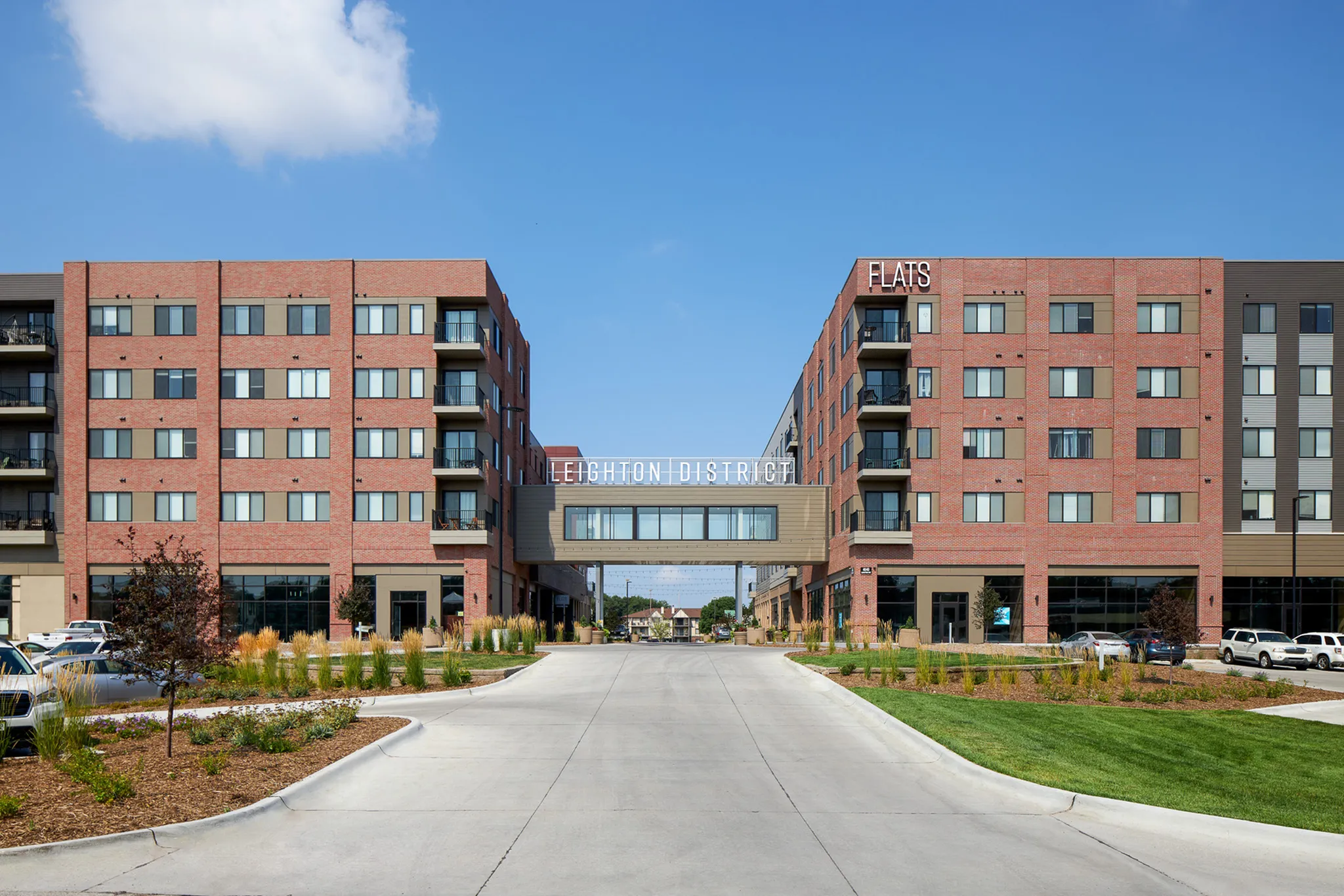 Modern apartment complex with red brick exterior, landscaped entrance, and bridge connection, featuring 'Leighton District' signage under a clear blue sky.