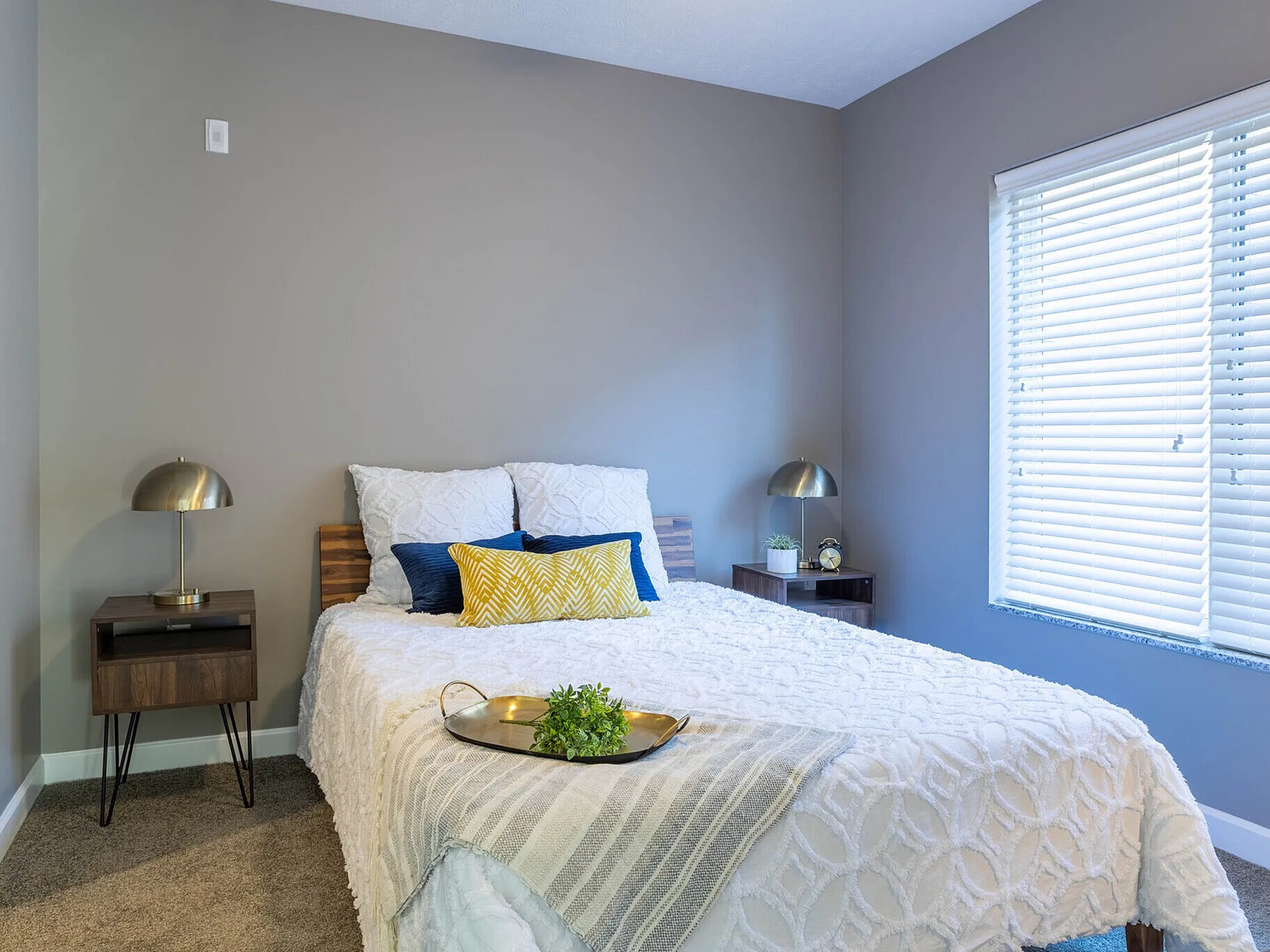 Inviting bedroom with cozy decor, featuring a plush white bedspread, modern lamps, and a sunlit window with blinds.