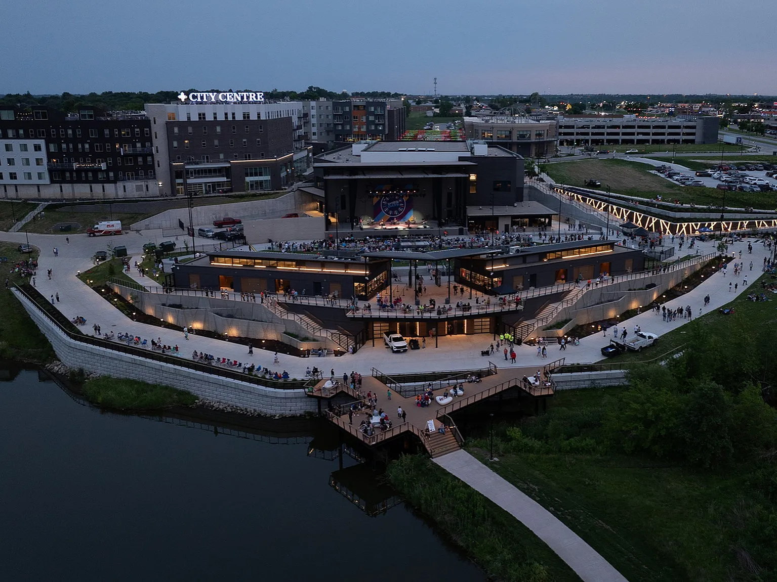 Vibrant evening view of City Centre with a bustling public event by the waterfront, showcasing modern urban architecture and lively community gathering spaces.