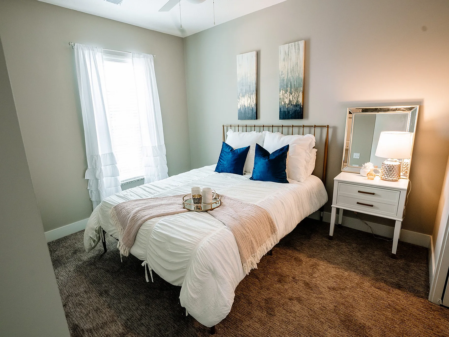 Cozy modern bedroom featuring a plush white bed with blue accent pillows, elegant nightstand with lamp, and stylish decor art above.