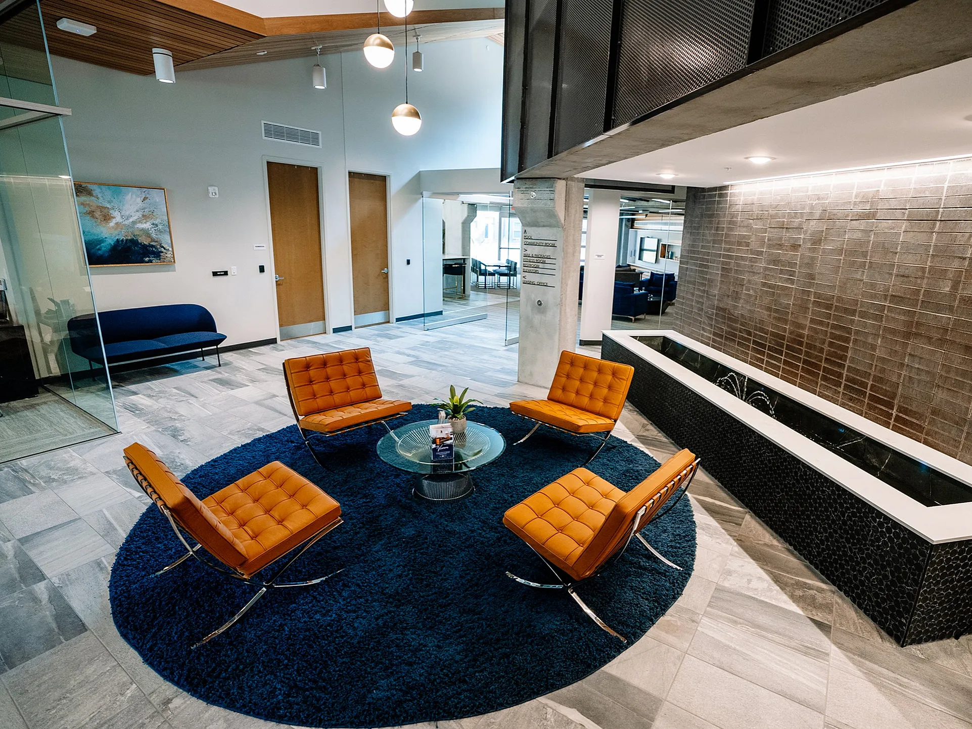 Contemporary lounge area with vibrant orange chairs, a sleek glass coffee table on a plush navy rug, and a stylish water feature under modern lighting.