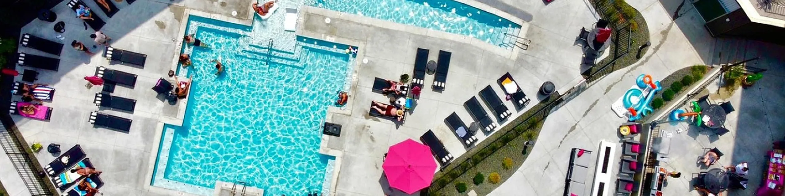 Aerial view of a vibrant apartment complex pool area featuring sparkling blue swimming pools, sun loungers, and pink umbrellas. Modern relaxation spaces and a lively atmosphere complete the luxurious setting.