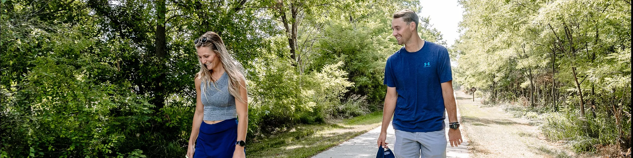 Joyful couple walking their dog on a scenic, tree-lined pathway surrounded by lush greenery.