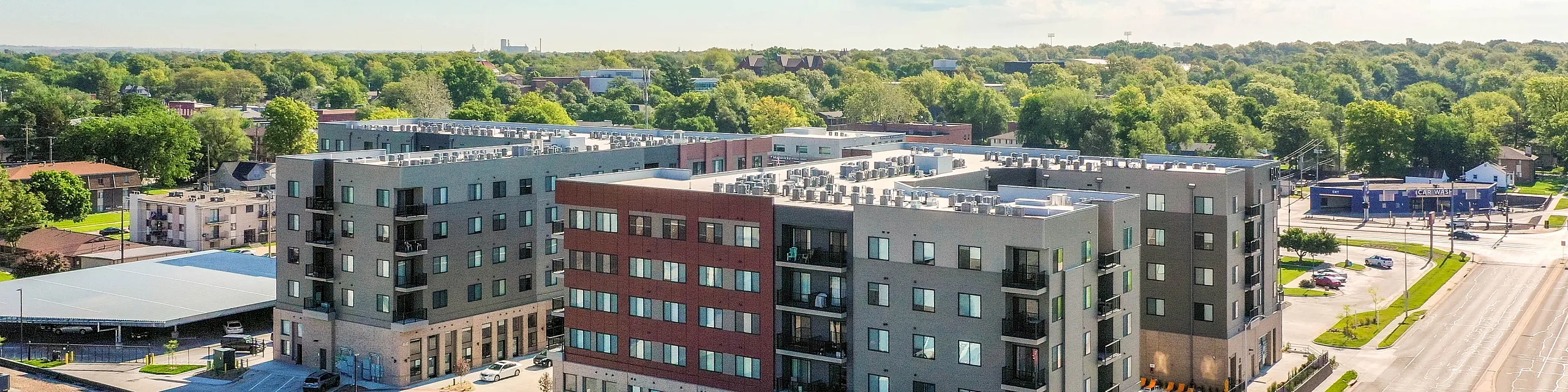 Modern apartment complex with sleek architecture, ample parking, and lush green surroundings under a bright blue sky.