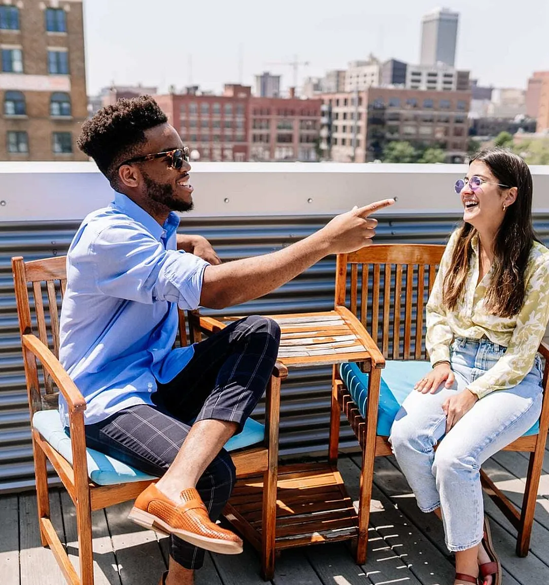 Vibrant rooftop patio featuring two people enjoying a conversation, surrounded by wooden chairs and an urban skyline backdrop.