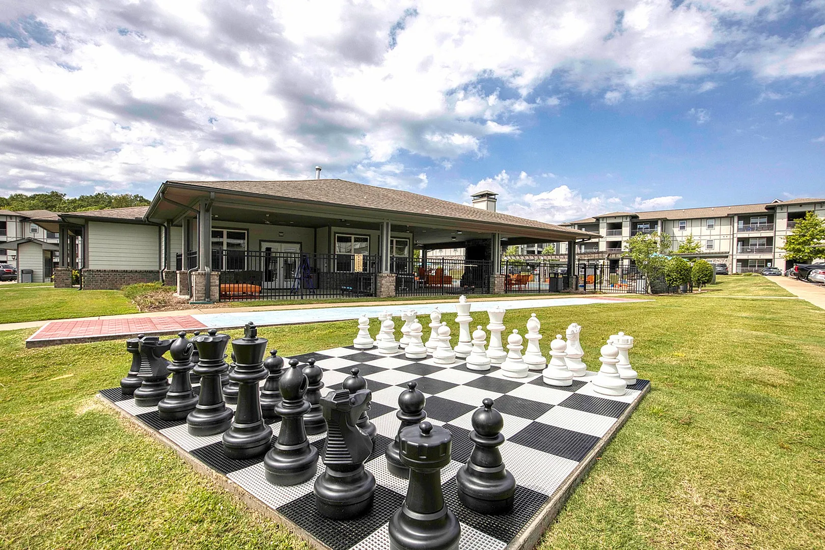 Vibrant apartment complex with a giant outdoor chess set, lush green lawn, and spacious community areas under a clear sky.