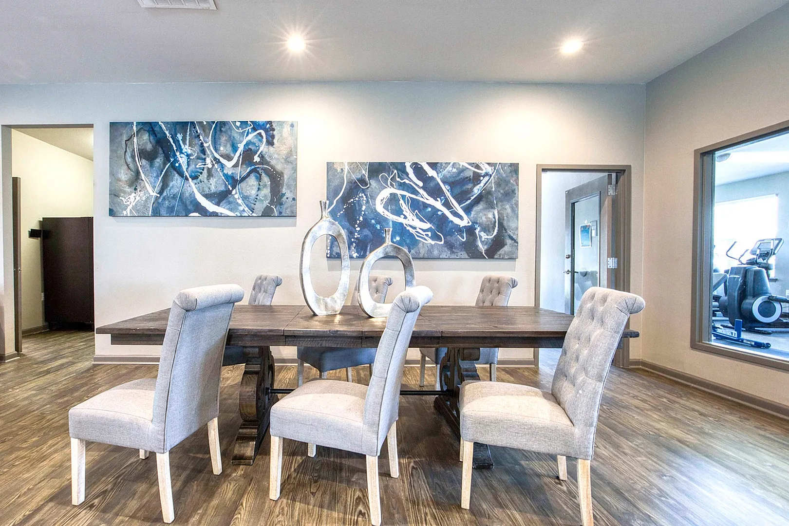 Elegant dining room with modern art, featuring a rustic wooden table and plush gray chairs, enhanced by contemporary sculptures and ambient lighting.
