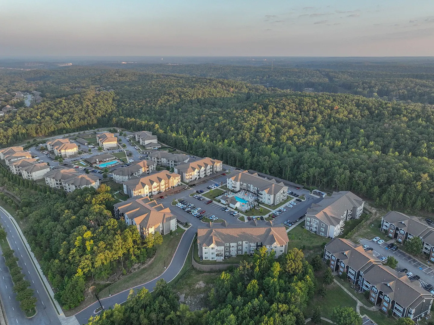 Aerial view of a modern apartment complex surrounded by lush greenery, featuring multiple buildings, ample parking, and a central swimming pool.