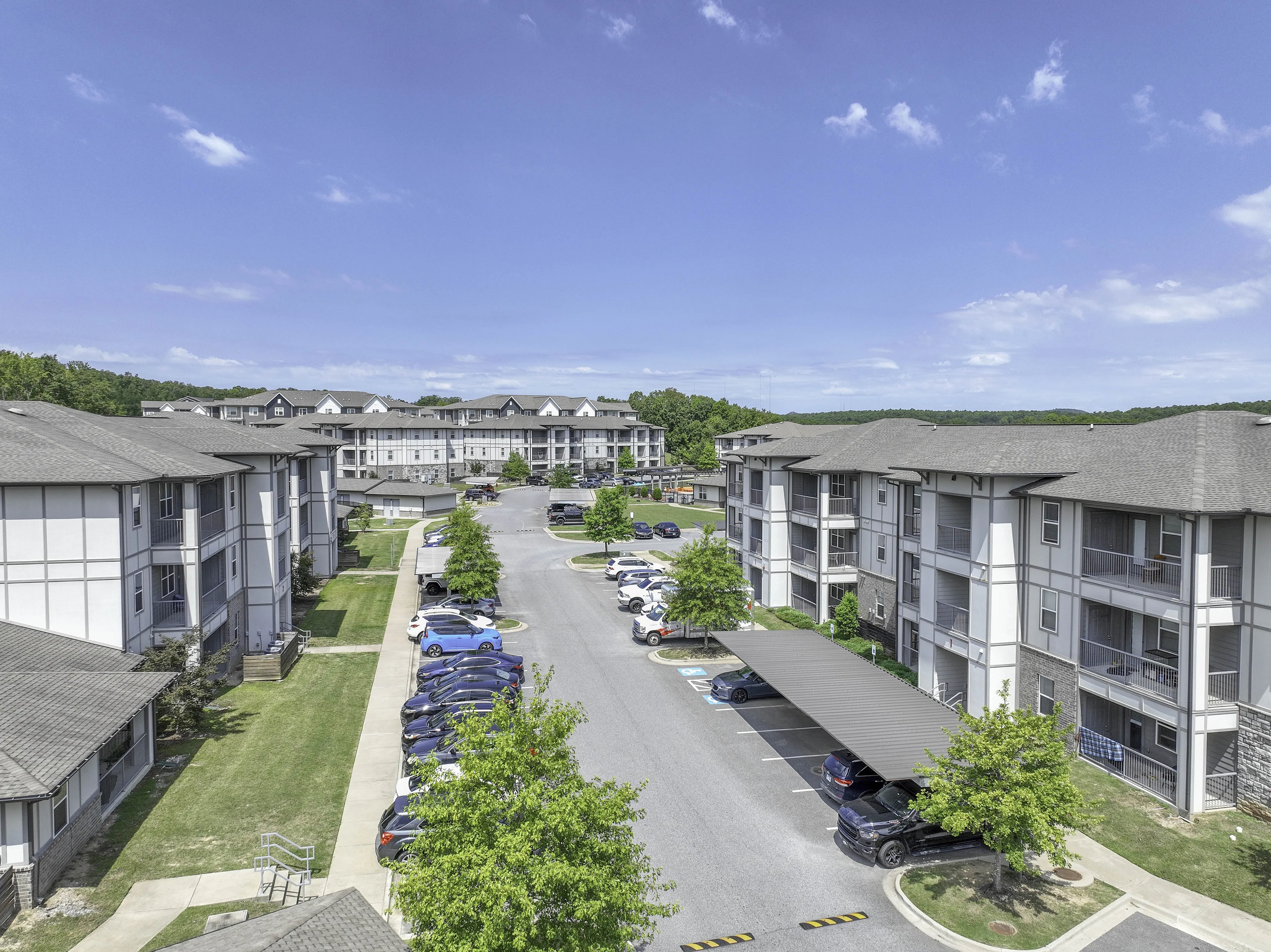 Sunny aerial view of a modern apartment complex with ample parking, lush greenery, and scenic surroundings.