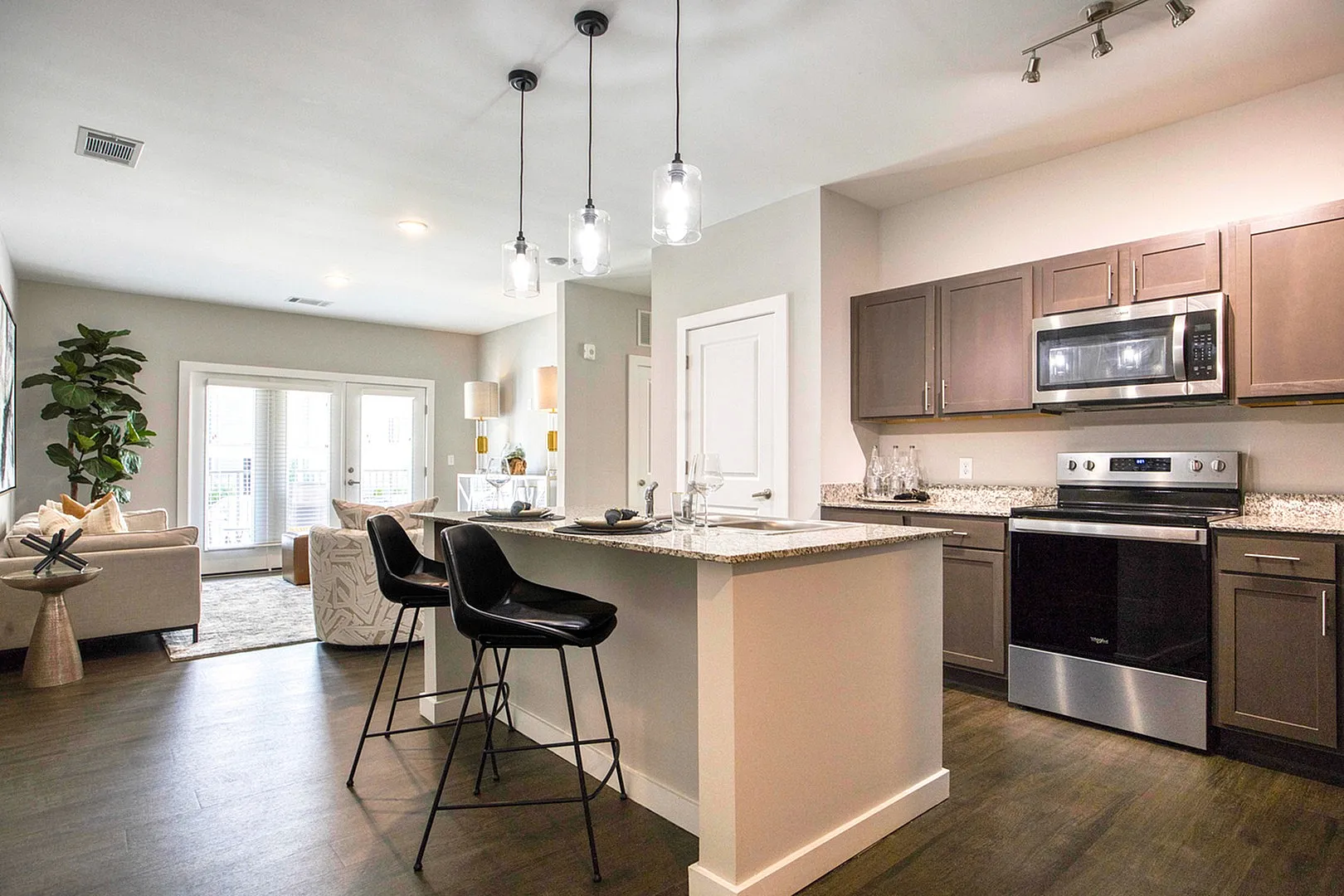 Modern open-plan kitchen and living area with sleek dark wood floors, stylish island counter, pendant lighting, and natural light flooding through large patio doors, accentuated by contemporary decor.