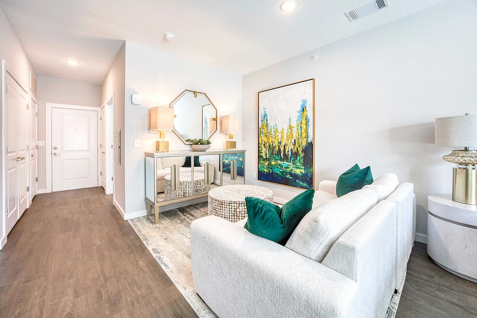 Elegant living room with tasteful decor, featuring a modern white couch with teal cushions, chic artwork, and a stylish mirrored console table under warm lighting.