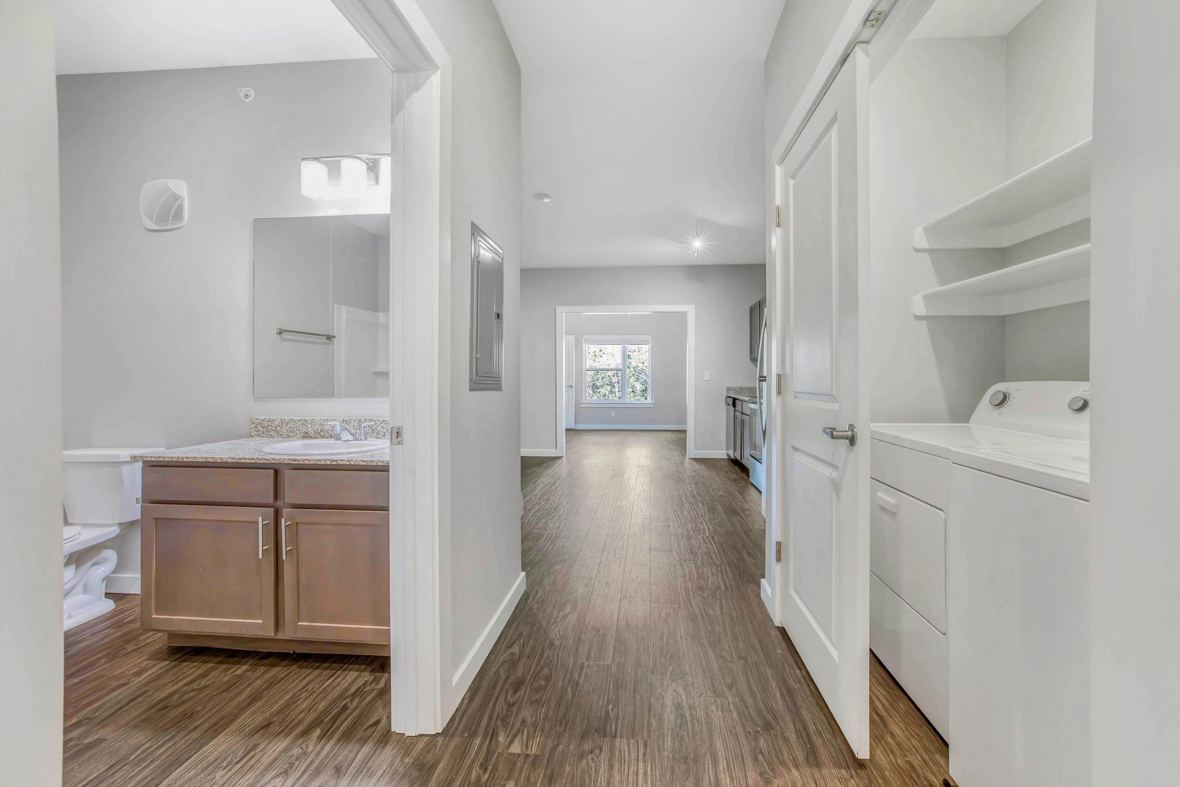 Modern apartment hallway featuring elegant wood flooring, clean lines, and natural light streaming in from large windows. Includes a cozy bathroom with granite countertops and an in-unit washer and dryer for convenience.