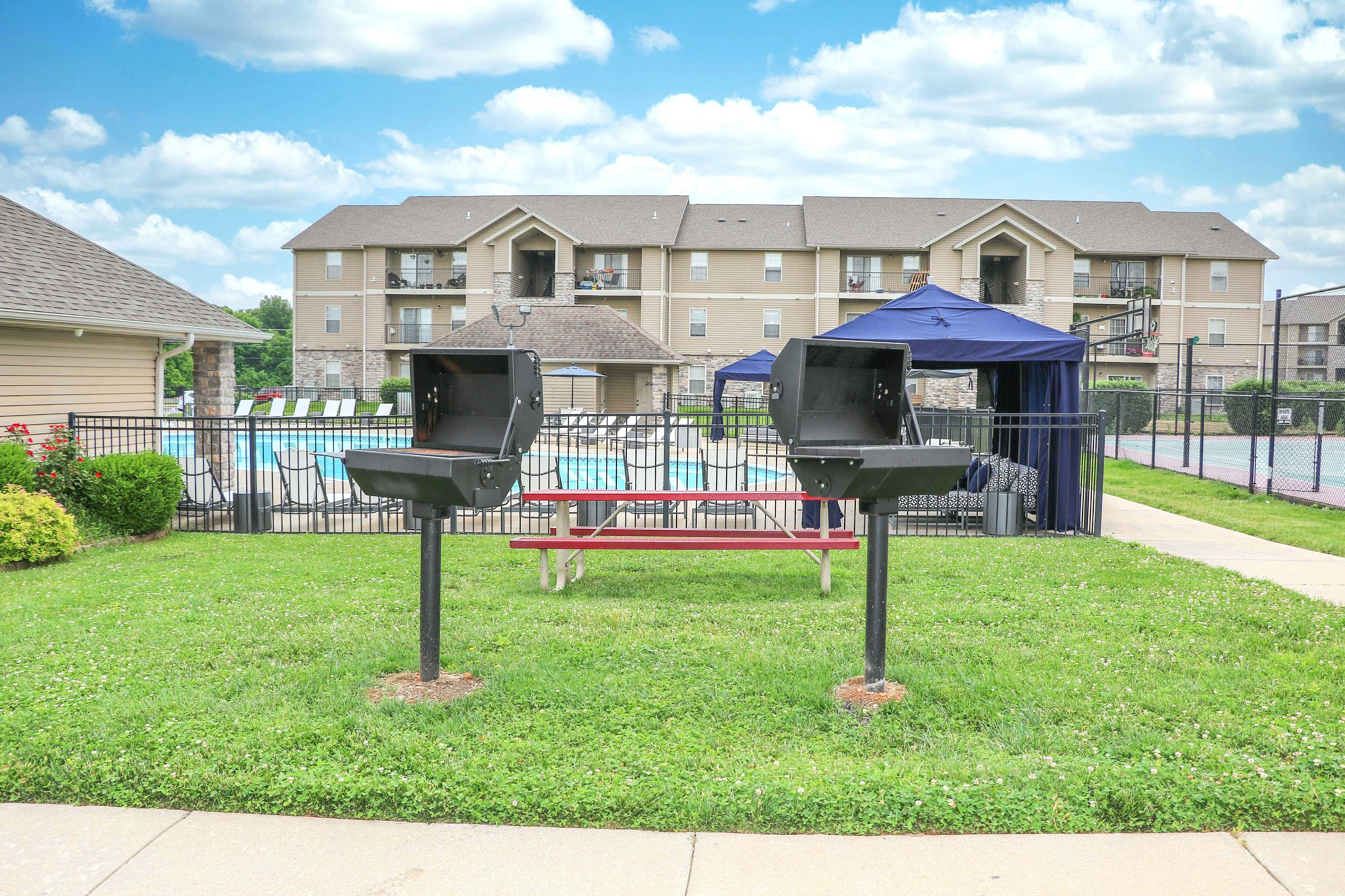 Inviting apartment complex with poolside relaxation area, barbecue grills, and sports courts under a clear sky.