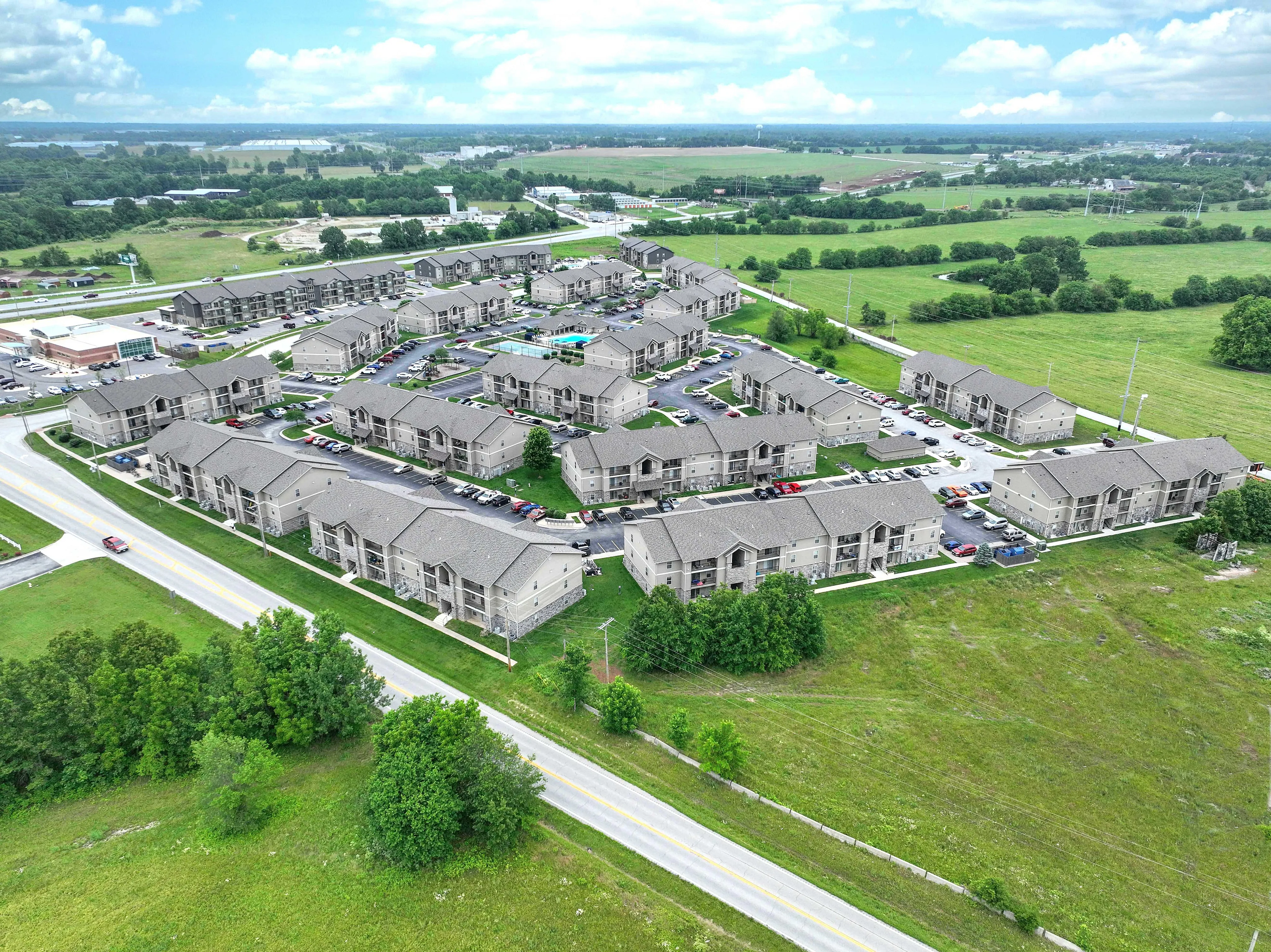 Aerial view of a modern apartment community surrounded by lush greenery, showcasing spacious buildings with abundant parking and scenic countryside vistas.
