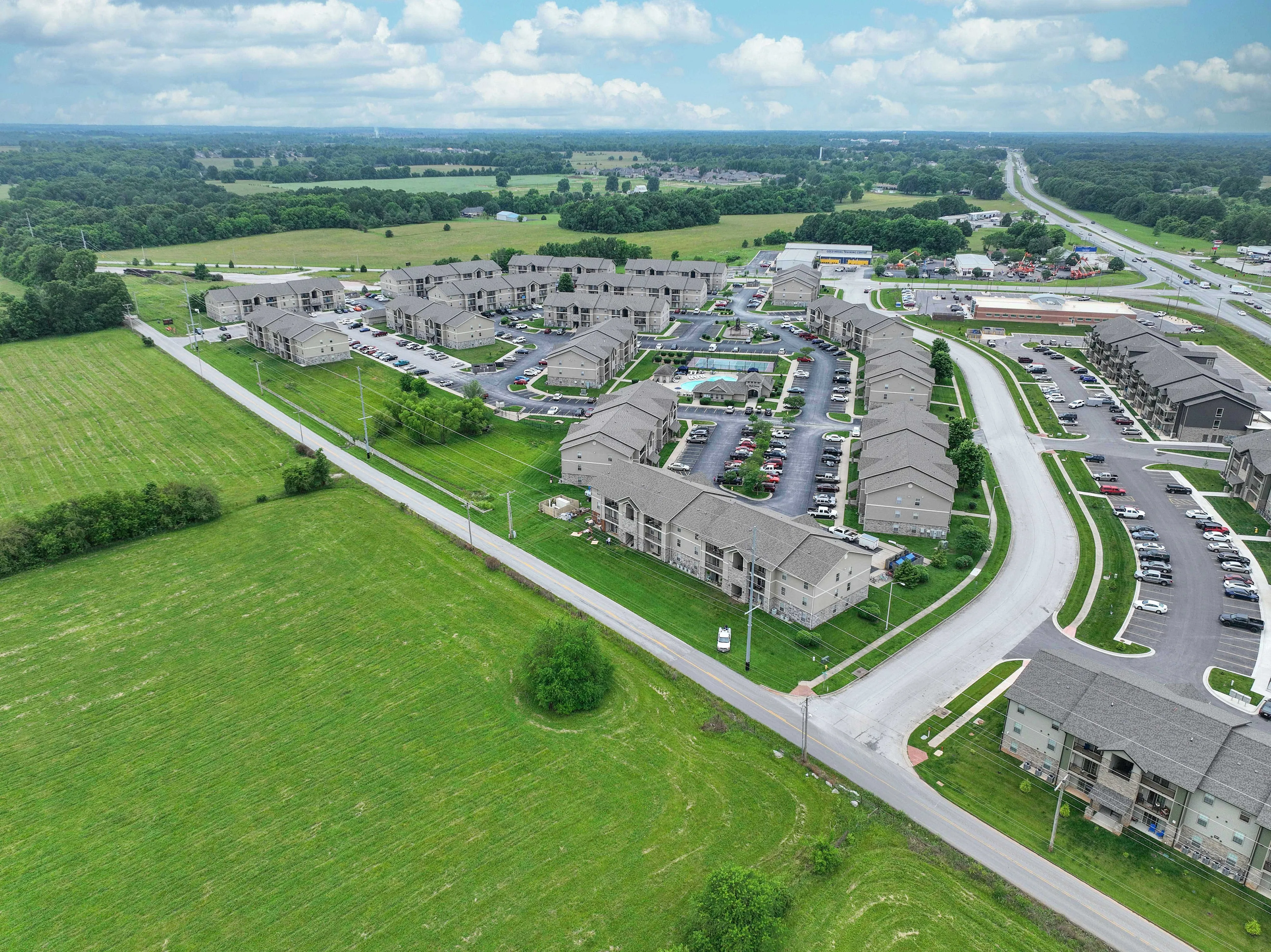 Charming aerial view of a modern apartment community, surrounded by lush greenery and spacious fields, showcasing convenient road access and ample parking.