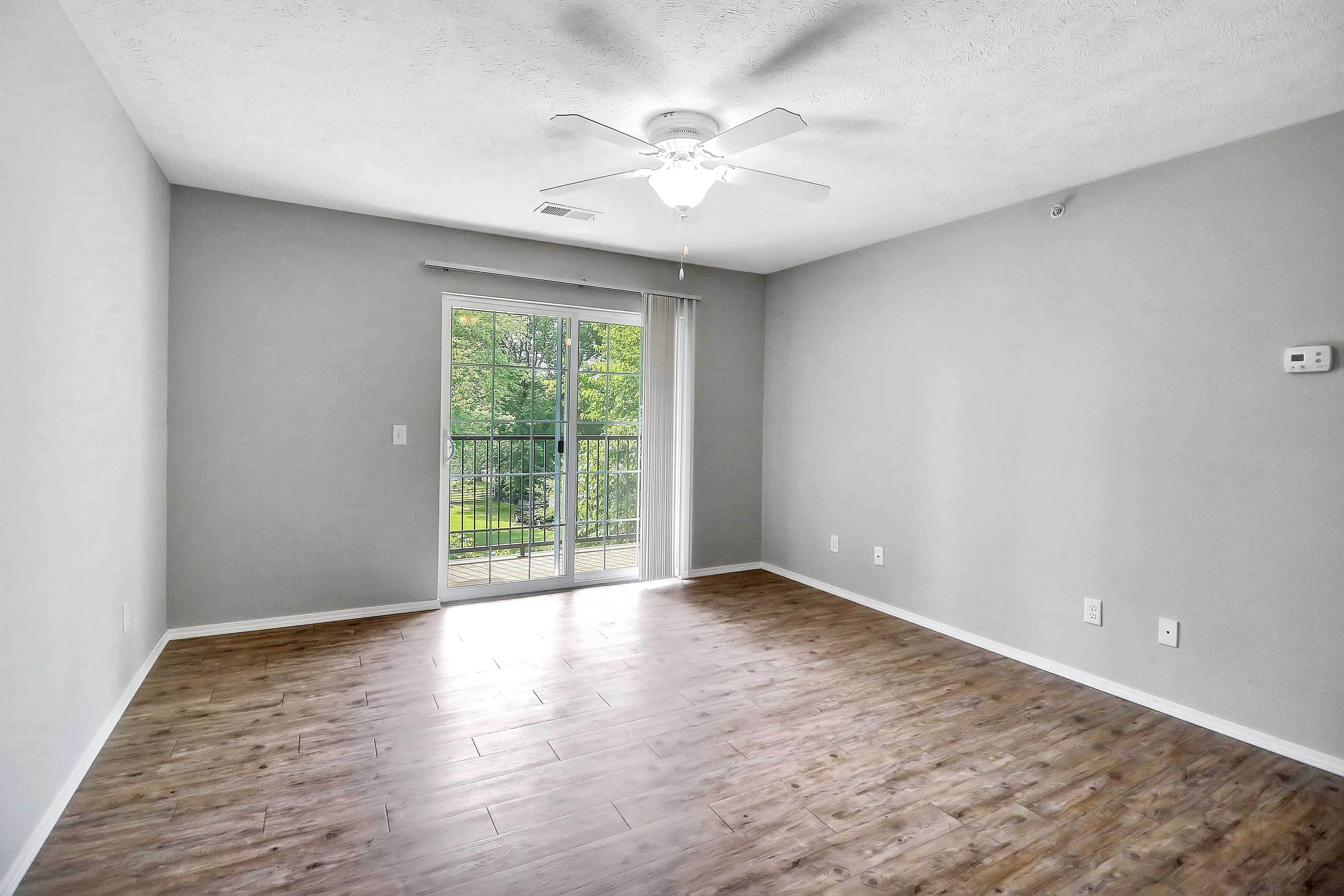 Bright and airy room with elegant wood flooring, ceiling fan, and a sliding door opening to a scenic balcony view.