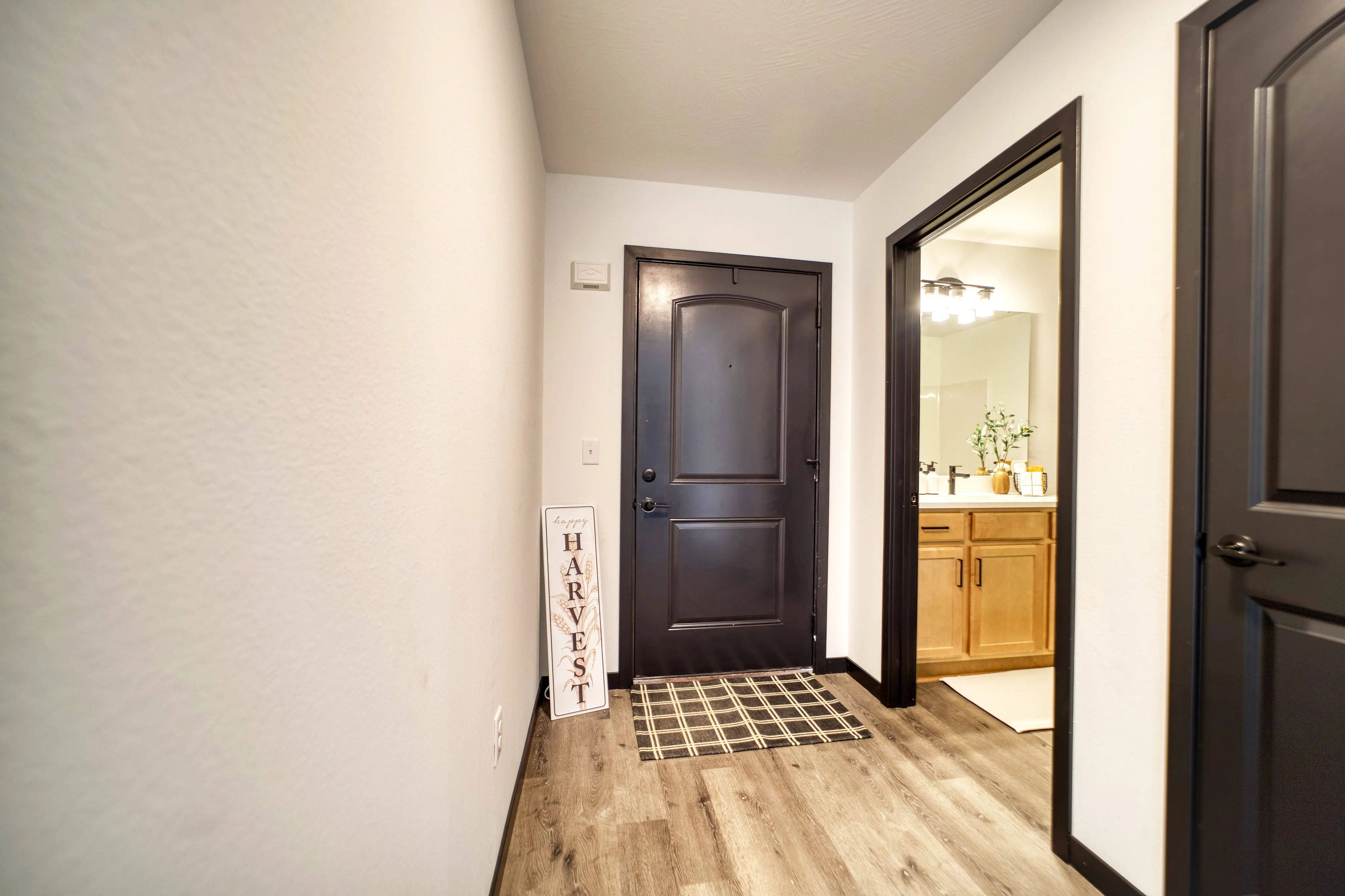 Welcoming apartment entrance featuring sleek dark doors, warm wood flooring, and a charming 'Happy Harvest' sign for a cozy aesthetic.