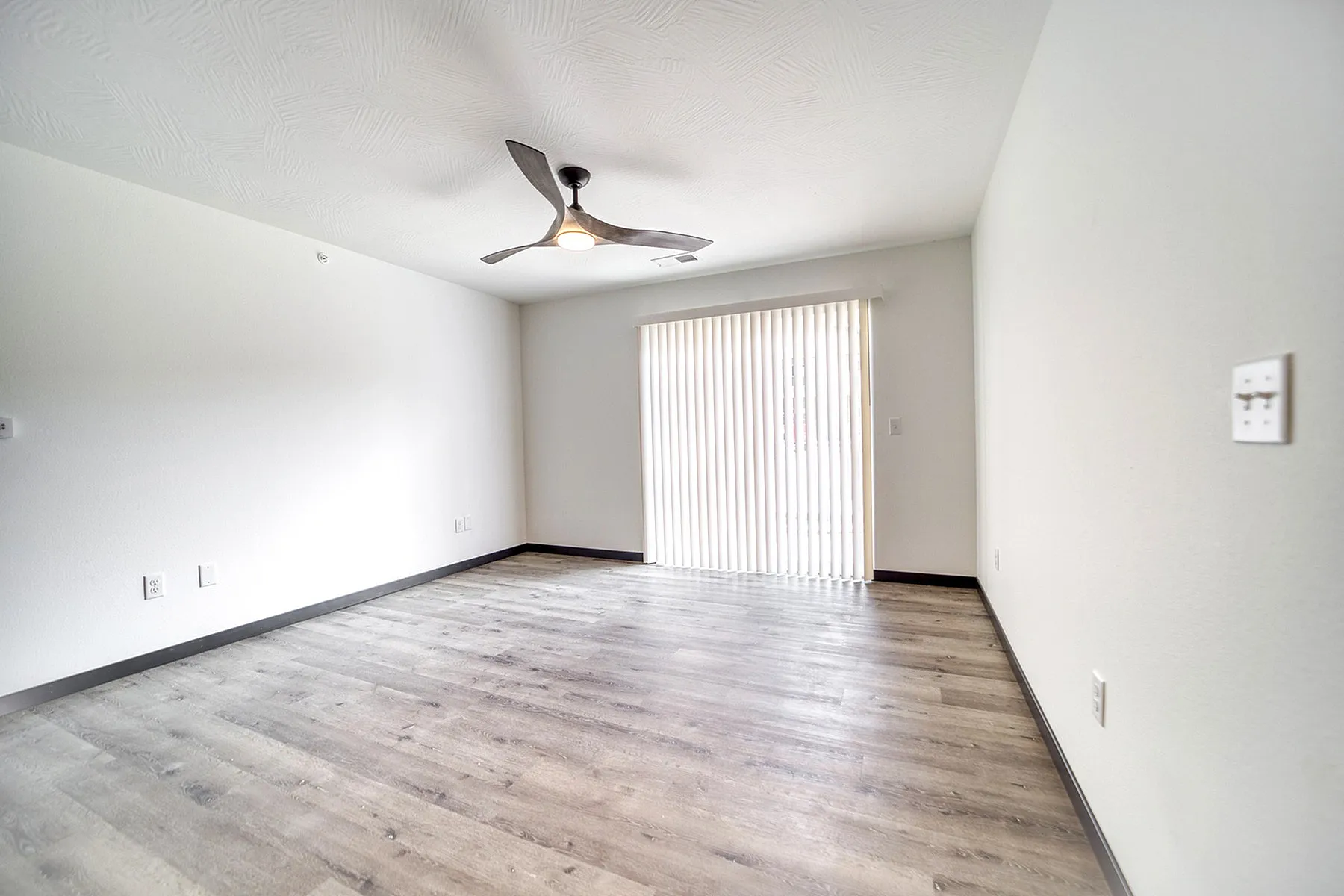 Bright and airy living space featuring modern vinyl flooring, sleek ceiling fan, and large sliding glass doors with vertical blinds.