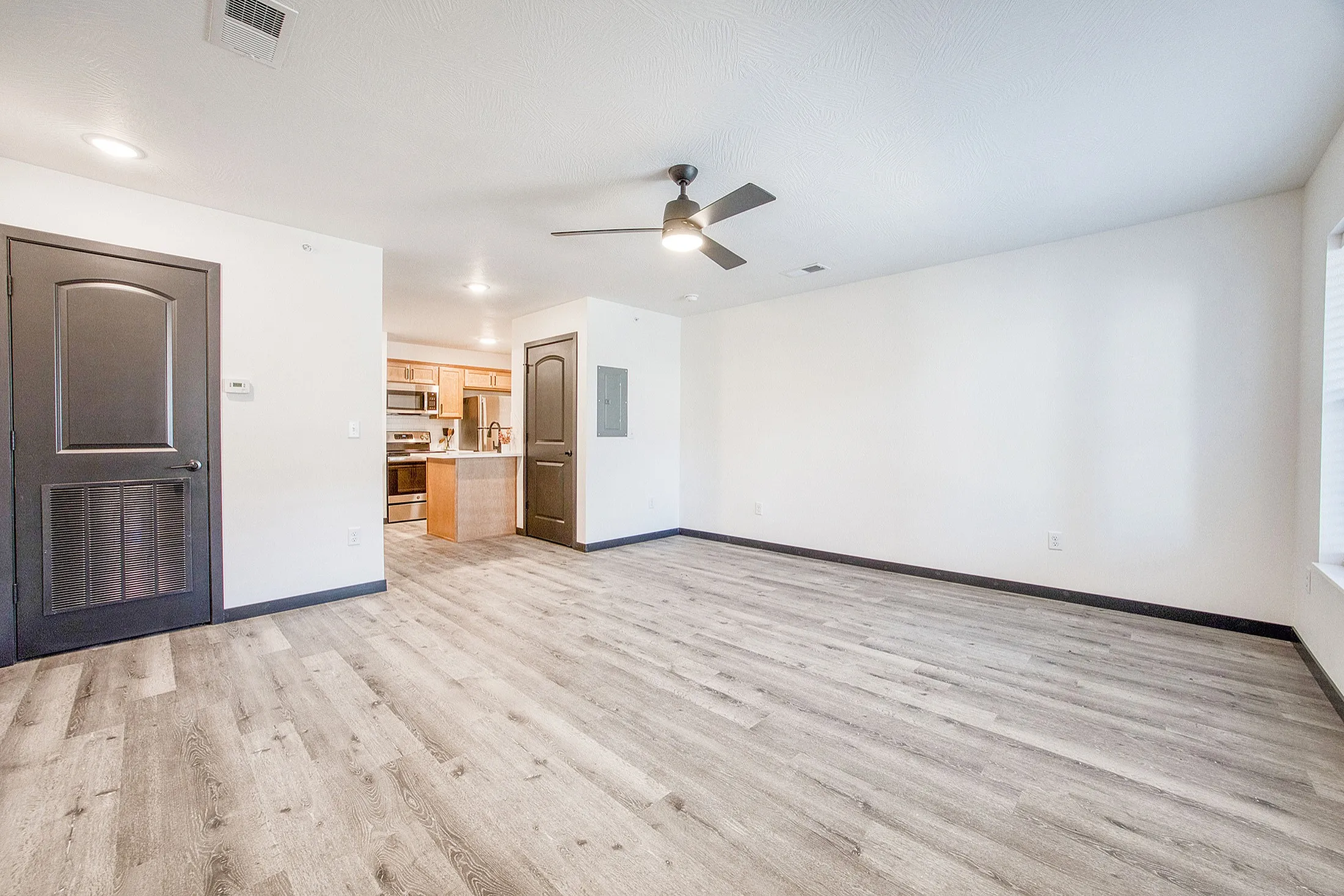 Sleek and modern living area with wood-inspired flooring, stylish ceiling fan, and open access to a contemporary kitchen with stainless steel appliances.