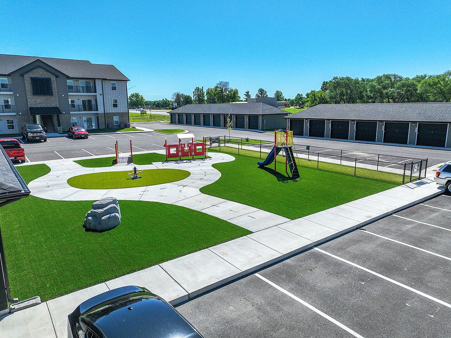 Modern apartment complex with a vibrant playground and ample parking, surrounded by lush greenery under a clear blue sky.
